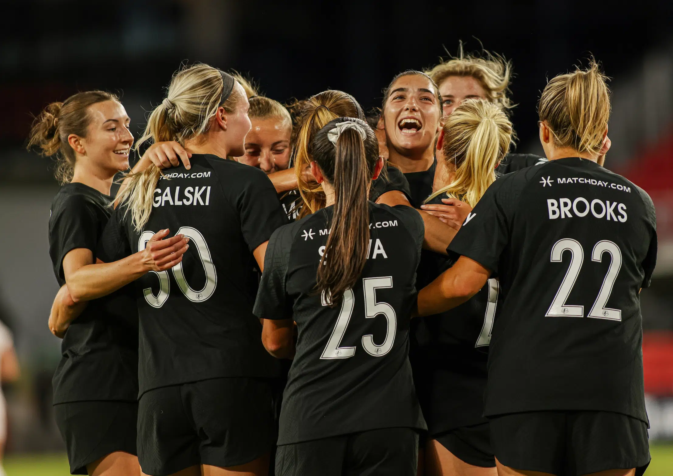 A group of soccer players wearing black jerseys hug and celebrate.