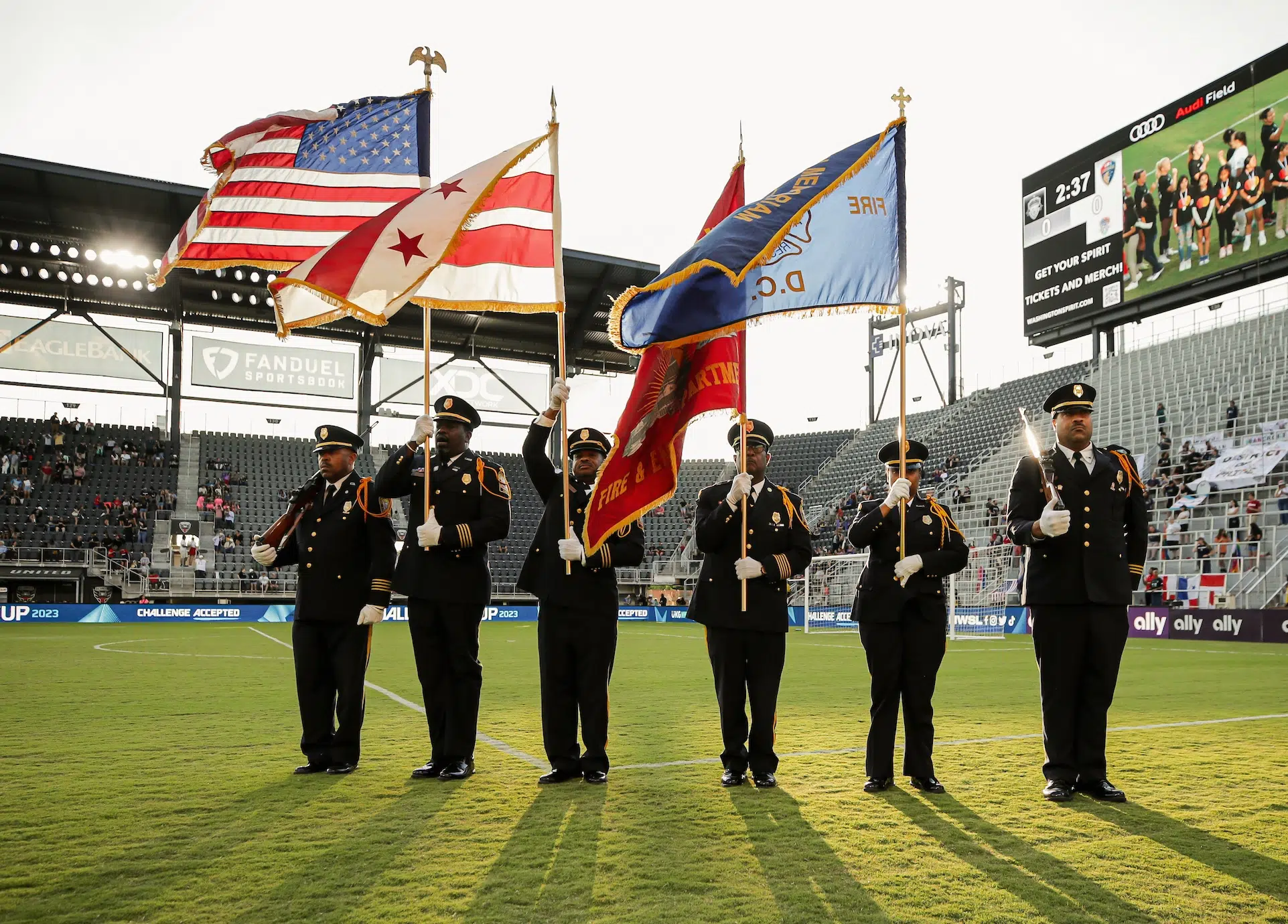 Six uniformed firefighters stand at attention and hold the American, DC, and DC Fire Department flags on a soccer field.
