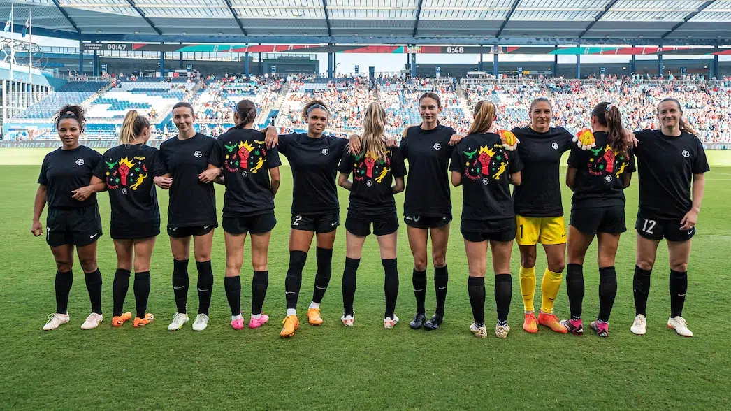 Eleven soccer players stand shoulder to should with every other one facing away. They're wearing black t-shirts with red fist and a yellow crown on the back.