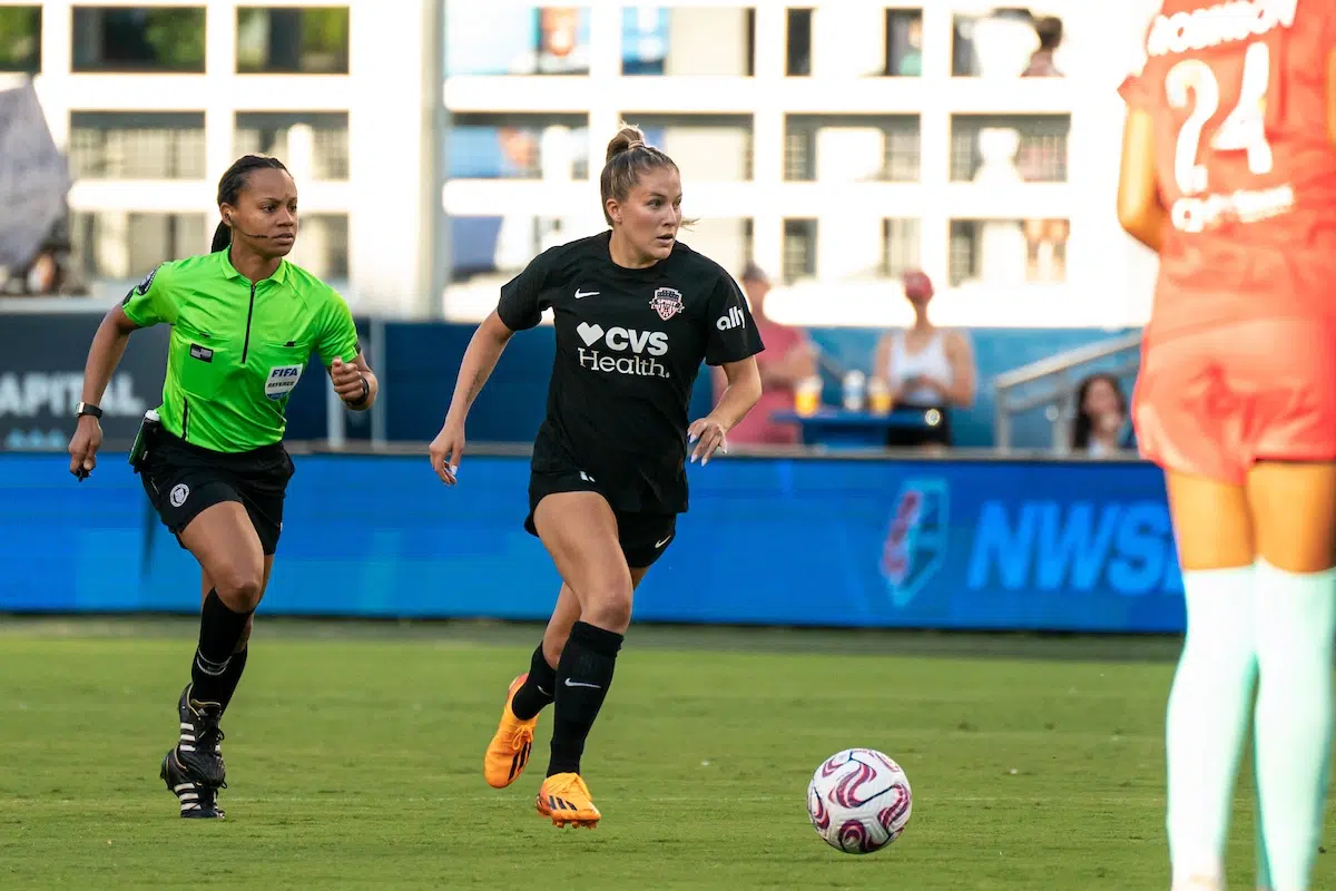 Ashley Sanchez in an all black uniform looks forward as she dribbles a soccer ball. A referee in a neon green shirt runs behind her.