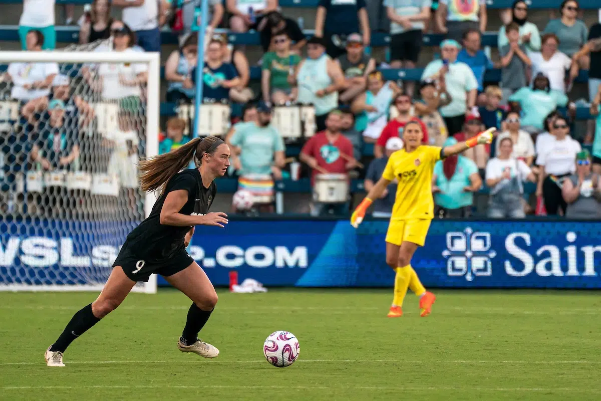 Tara McKeown in a black uniform dribbles a soccer ball. In the background, Aubrey Kingsbury in an all yellow uniform points her finger off to her left.