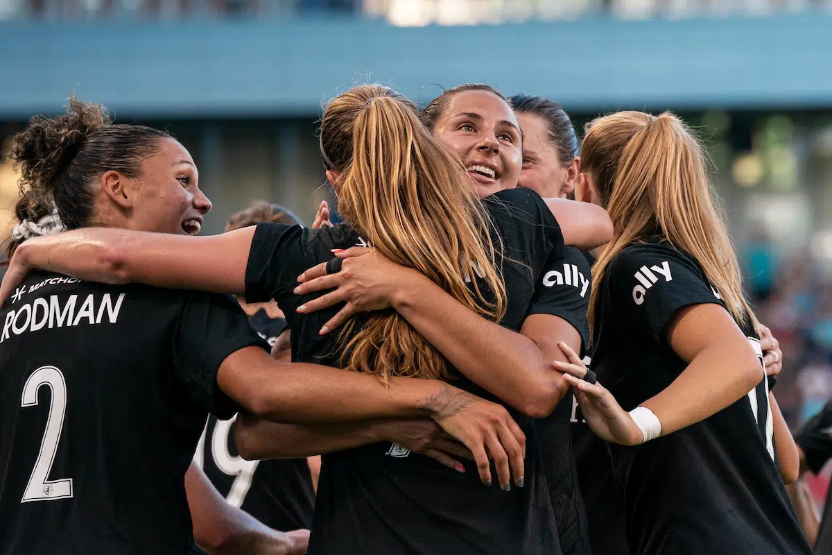 A group of soccer players in black jerseys hug after scoring a goal.