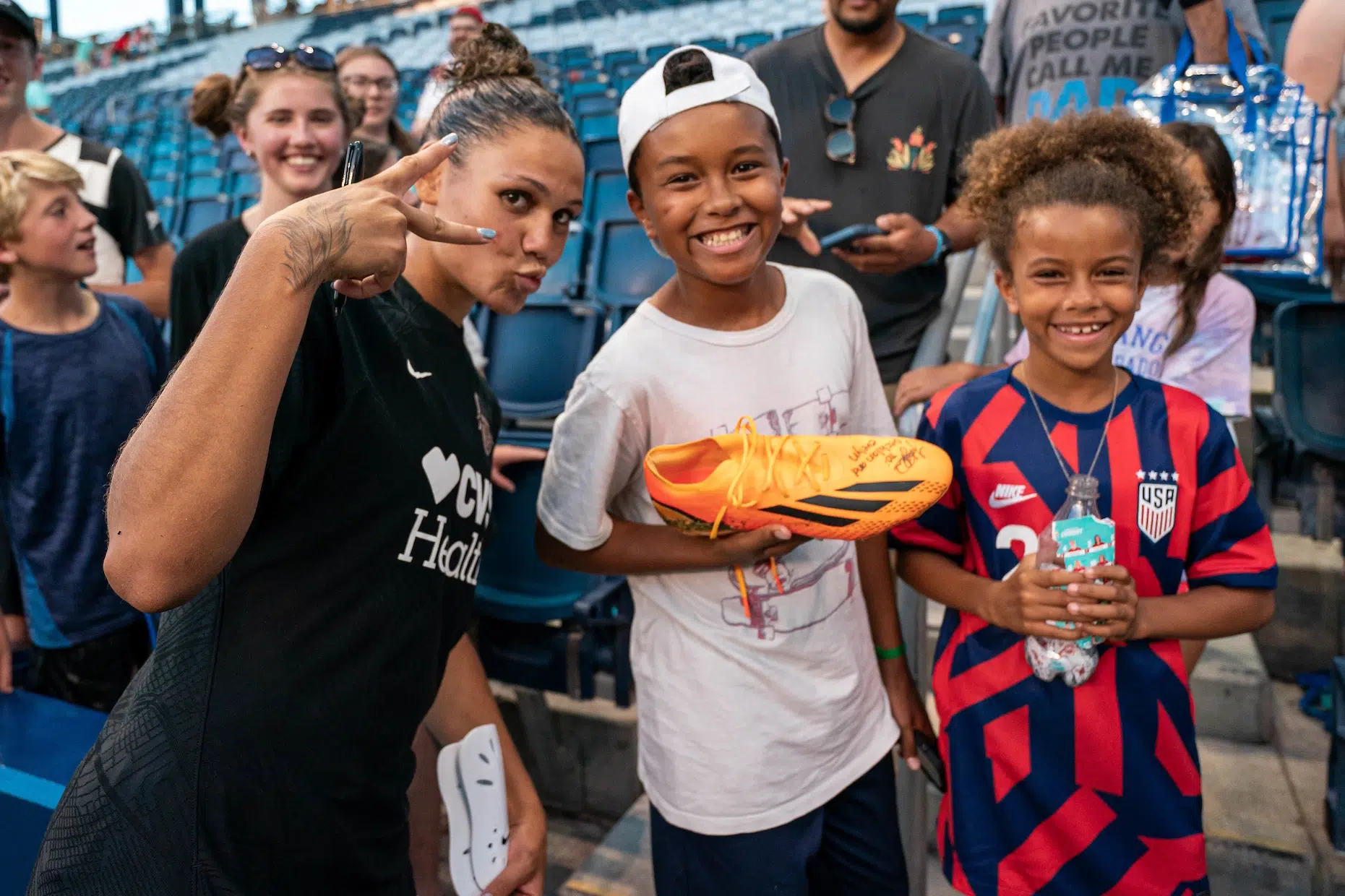 Trinity Rodman in a black jersey holds up a peace sign as she poses for a photo with two young fans. The fan closest to her holds one of her orange cleats with her autograph.