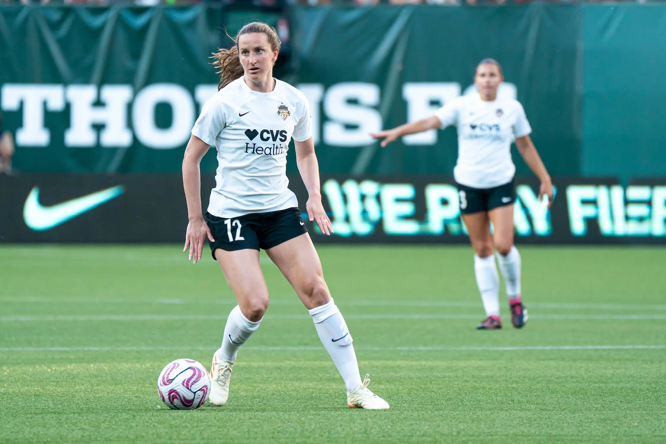 Andi Sullivan surveys the field while dribbling a soccer. She is wearing a white top, black shorts, and white socks.