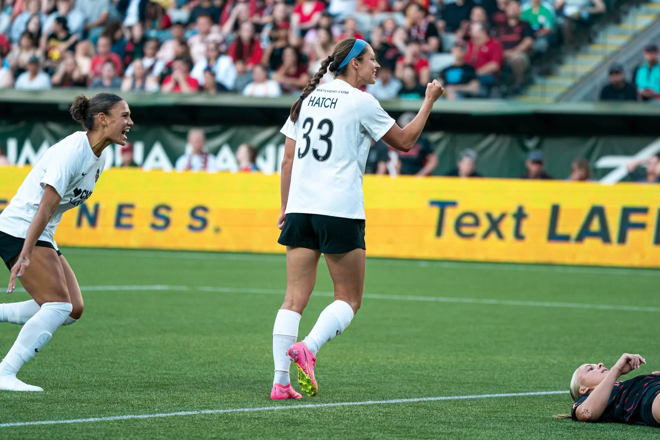 Ashley Hatch in a white top, black shorts and pink cleats pumps her fist in celebration as Trinity Rodman cheers and runs towards her.