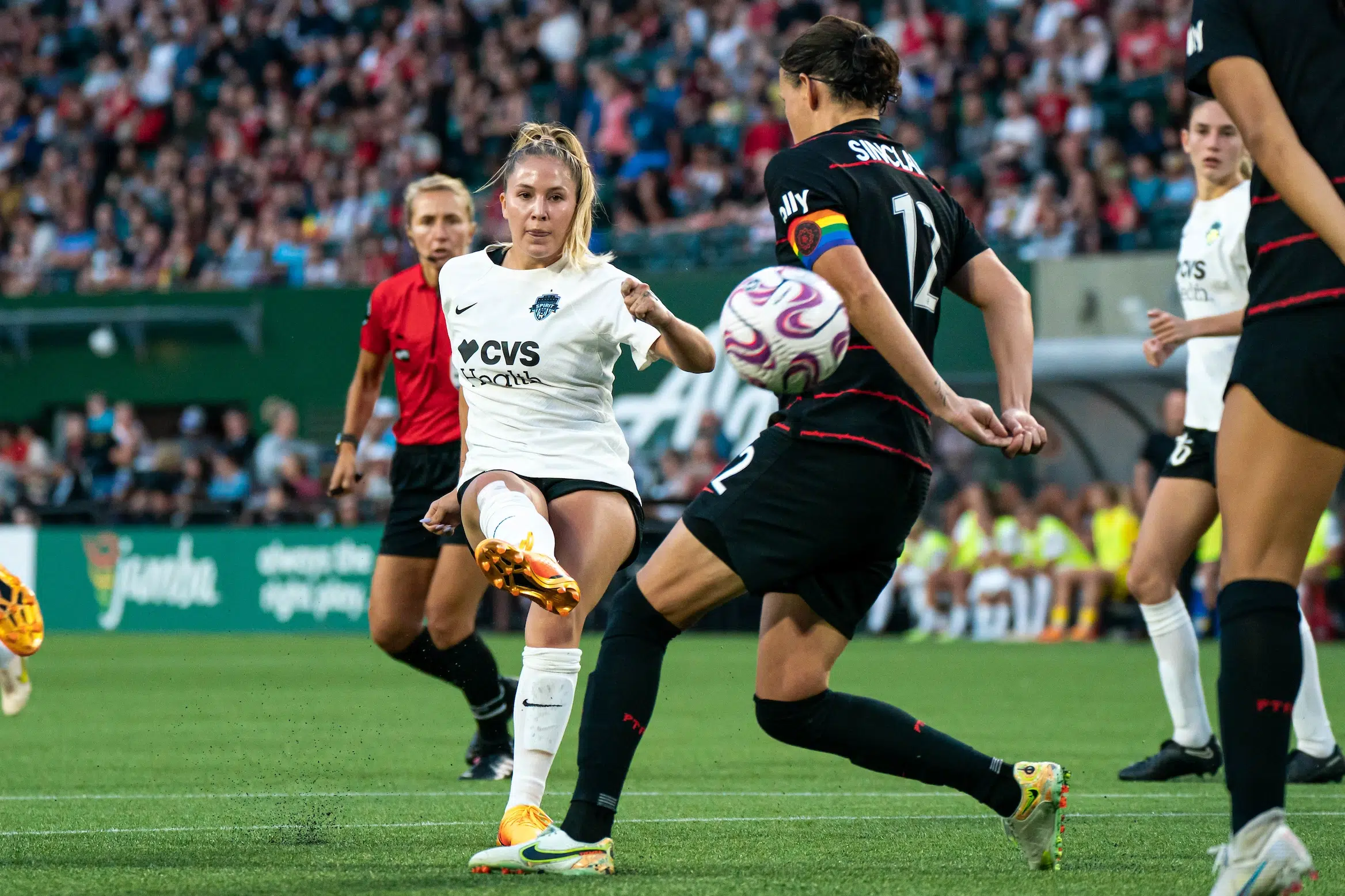 Ashley Sanchez in a white top, black shorts and white socks shoots the ball passed Christine Sinclair in a black uniform.