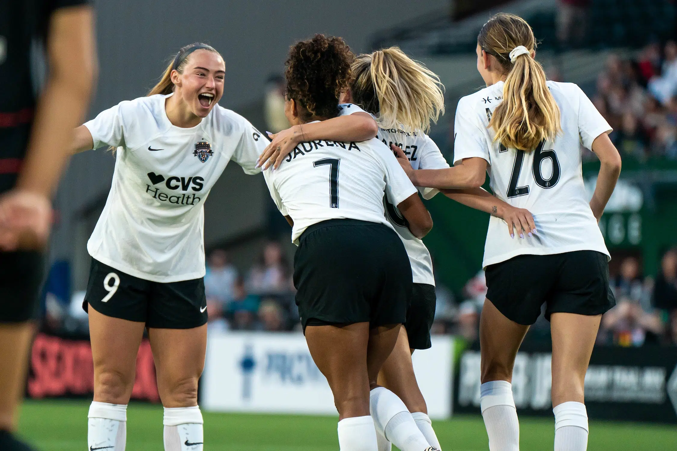 Four players in white jerseys, black shorts and white socks embrace and cheer after scoring a goal.