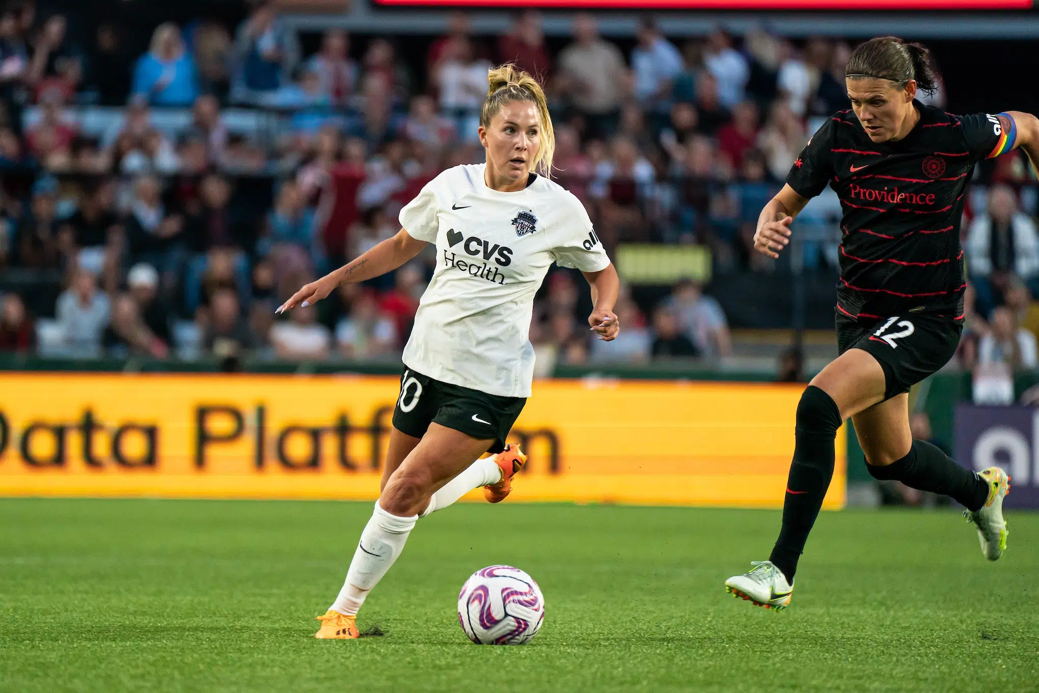Ashley Sanchez in a white jersey, black shorts and white socks dribbles the ball past Christine Sinclair who is wearing a black uniform.
