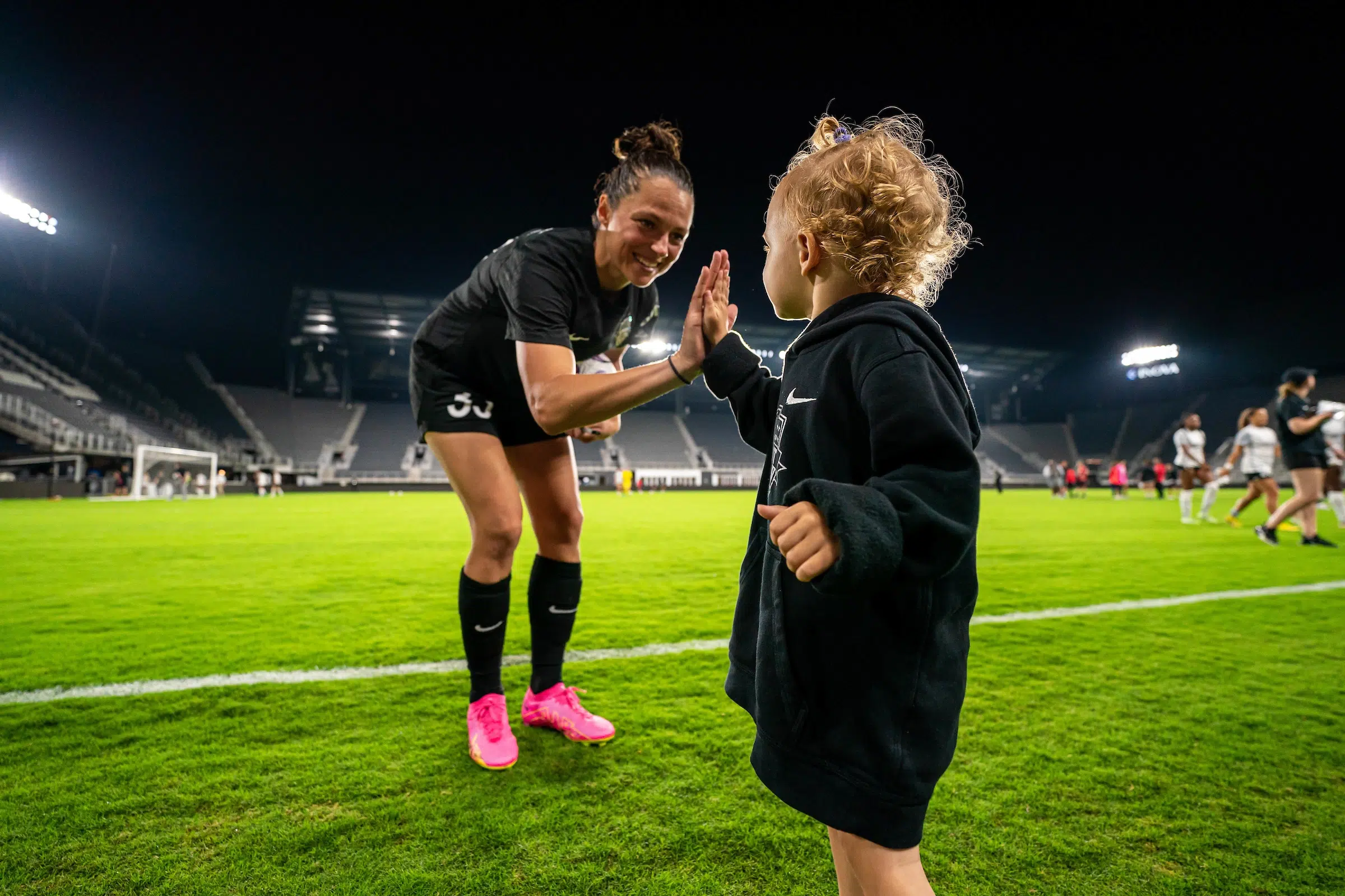 Ashley Hatch in a black uniform and pink cleats bends down to high five a toddler fan who is wearing a black hoodie.