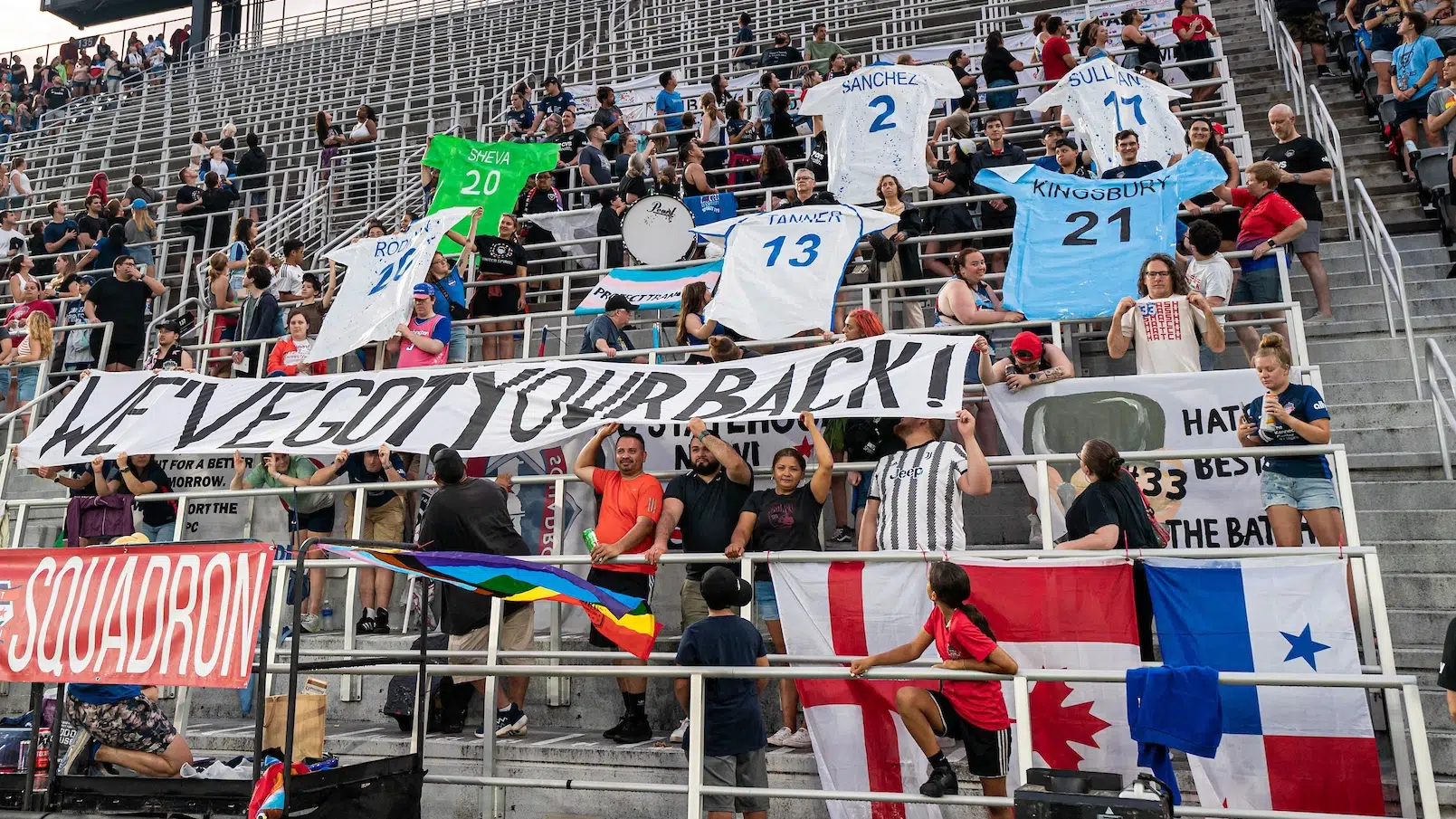 Fans hold up giant posters of jerseys for six players headed to the World Cup. A banner reading "We've Got Your Back" is held up by fans in the first row.