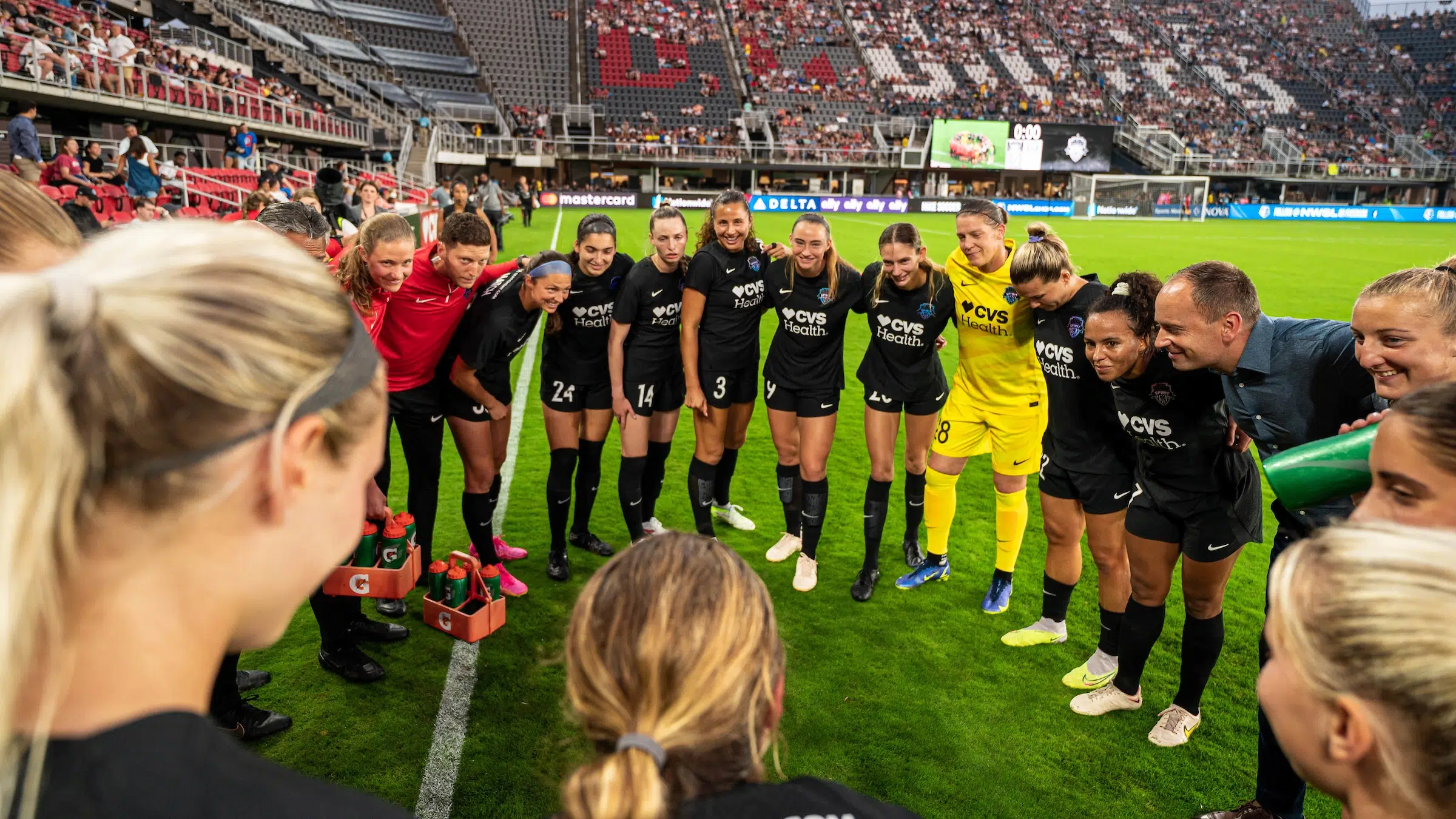 A soccer team in black uniforms huddles up alongside coaches and trainers in red tops.