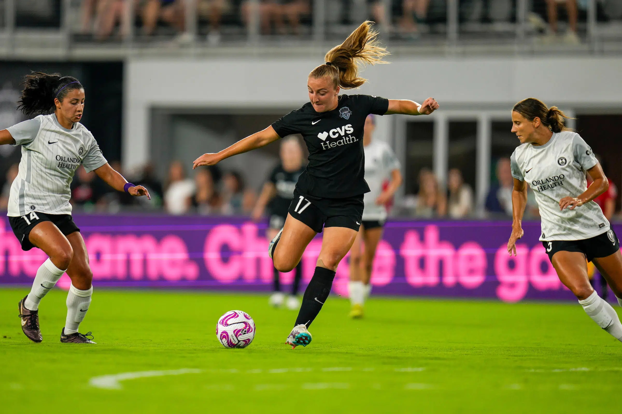 Nicole Douglas in an all black uniform dribbles the ball between two defenders in gray shirts and black shorts.