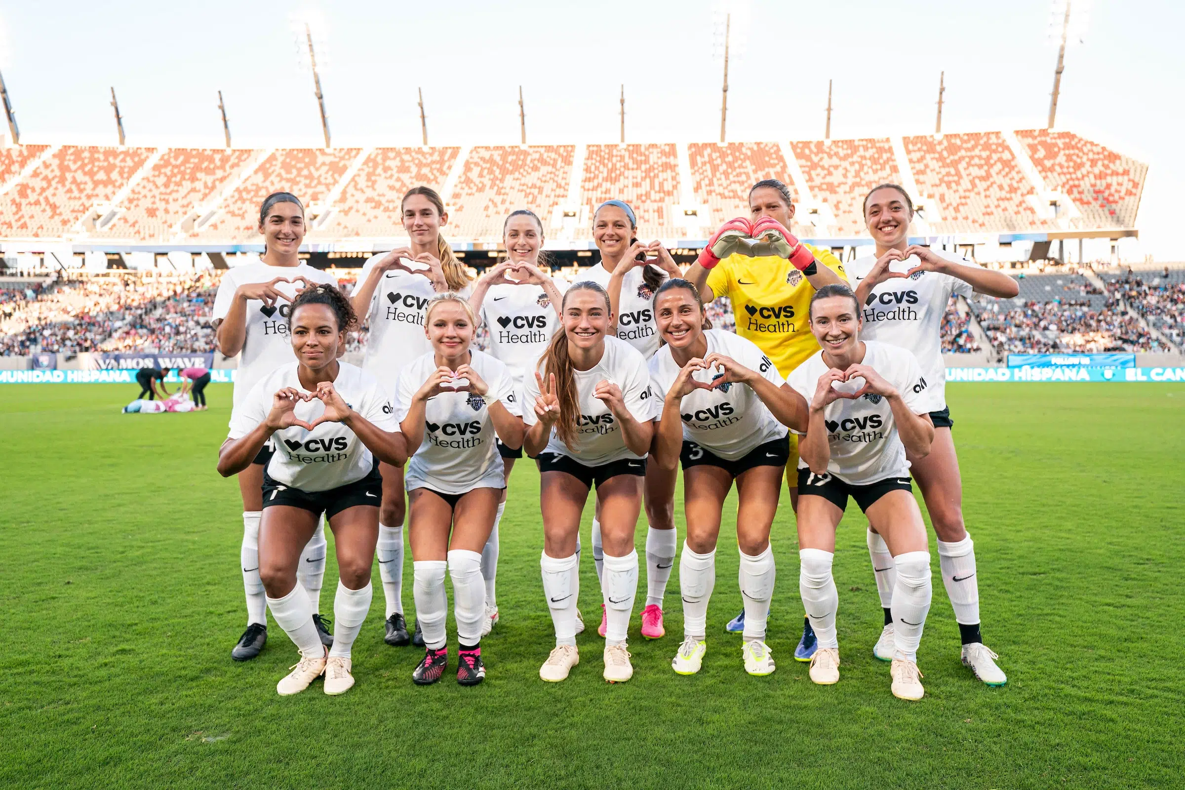 Ten soccer players in white uniforms and one in a yellow uniform pose with their hands making hearts.
