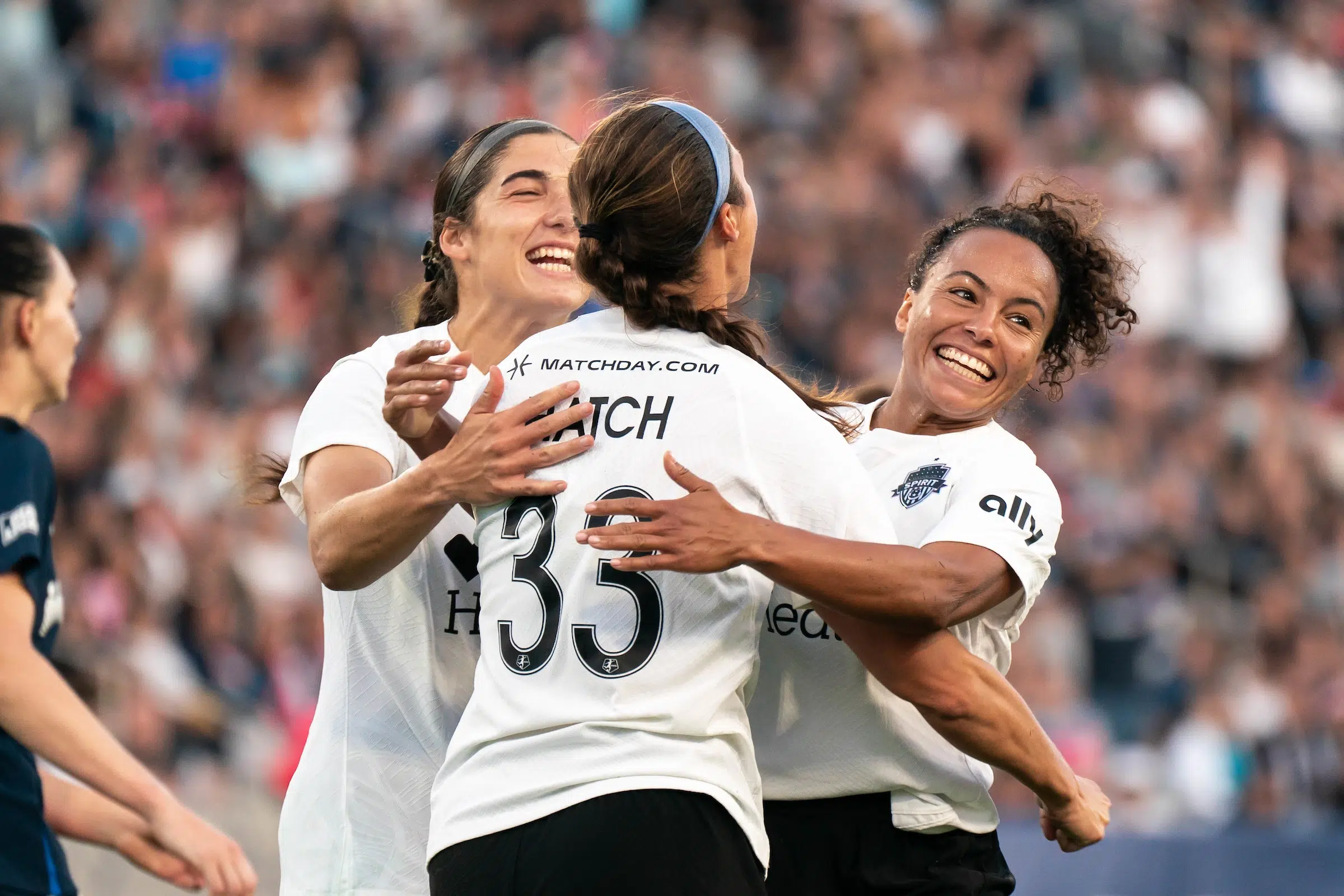 Three soccer players in white tops embrace and smile in celebration on a soccer field.