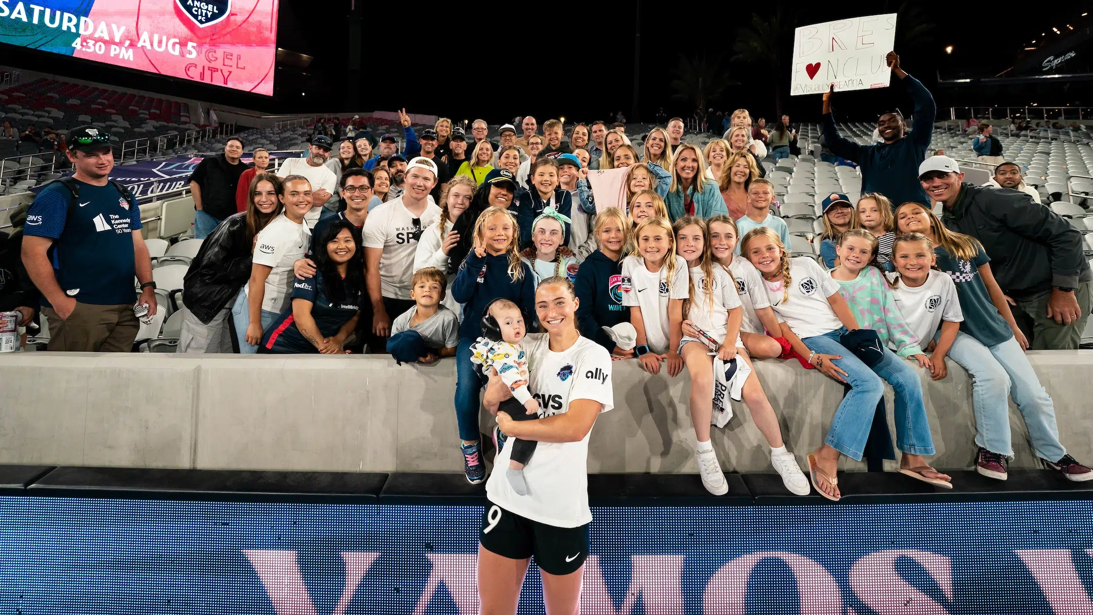 Tara McKeown in a white top and black shorts holds a baby wearing ear months in front of a group of fans filled into the stands at a soccer stadium.
