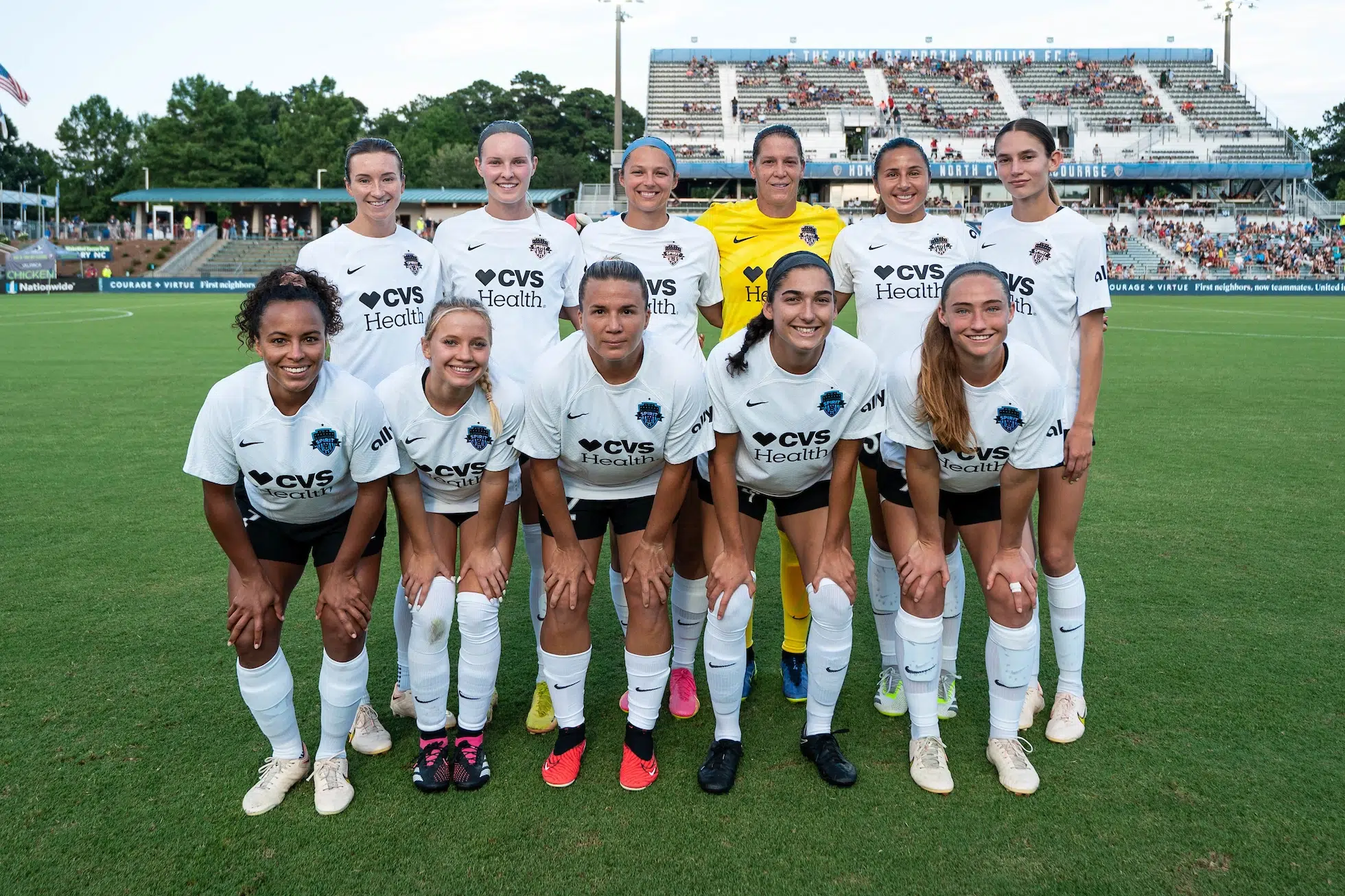 Ten soccer players in white uniforms and one in a yellow uniform pose for a photo in two rows.
