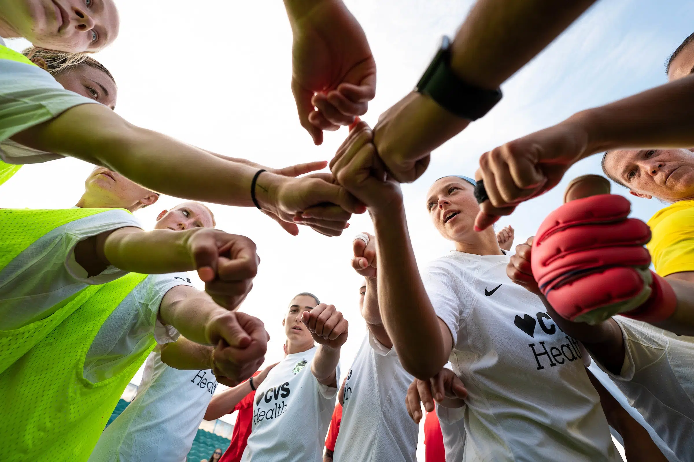A group of soccer players in white uniforms huddle up and put their fists in the middle to cheer.