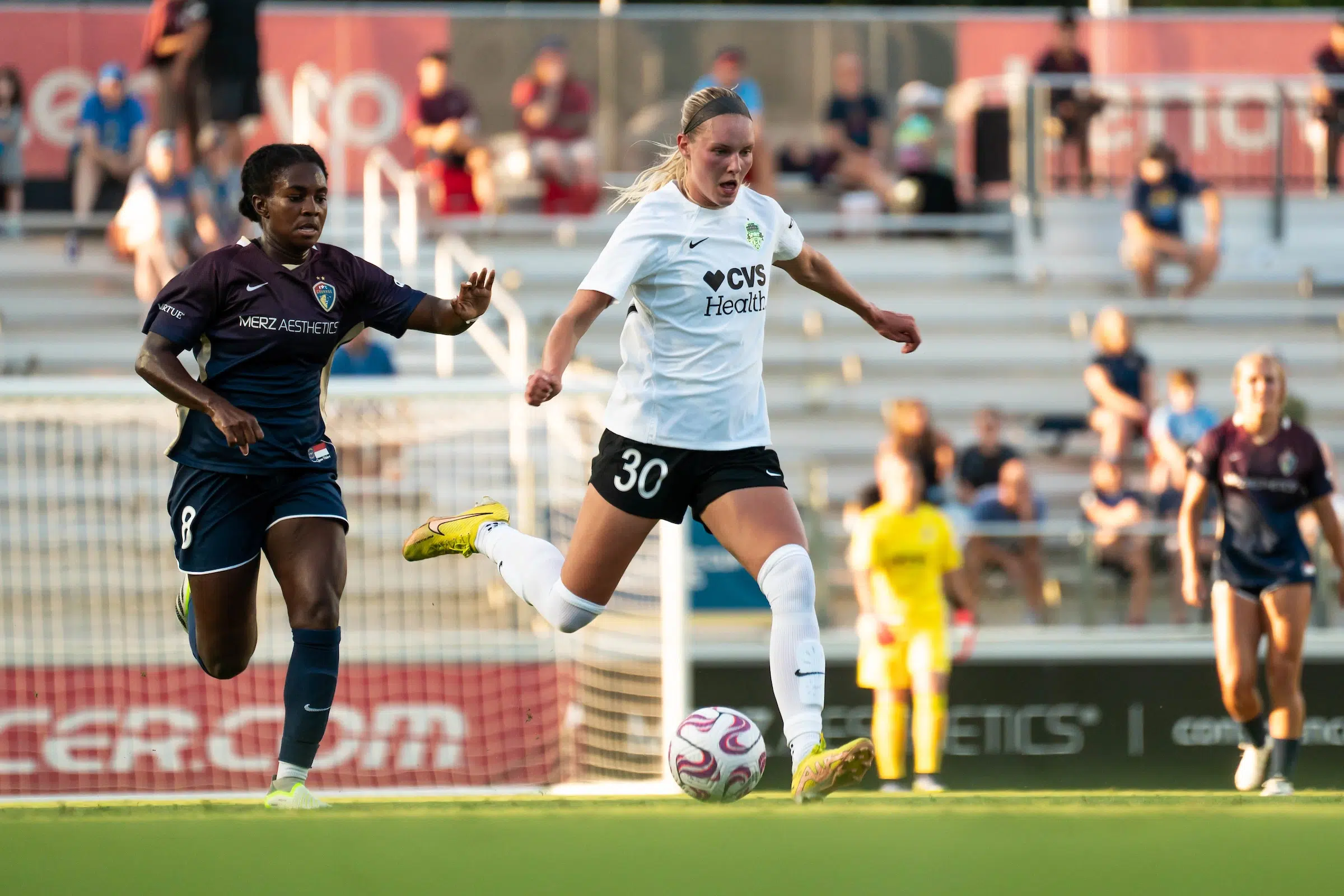 A soccer player in a white top, black shorts and white socks goes to kick a ball as a defender tries to chase her down on the soccer field.
