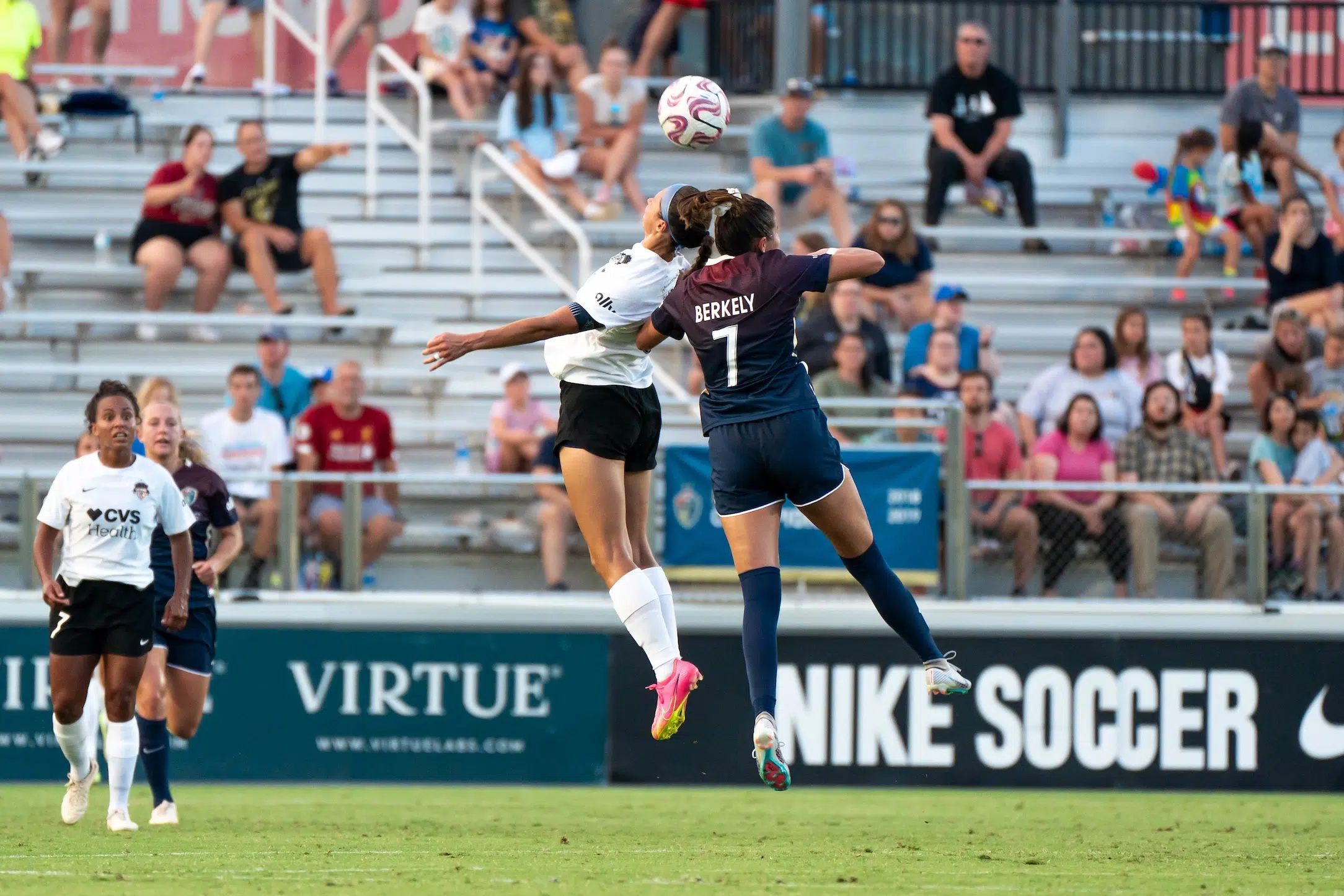 Ashley Hatch in a white uniforms battles for a header against a defender in a maroon to navy ombre uniform. The ball is approaching the two players who have jumped into the air.