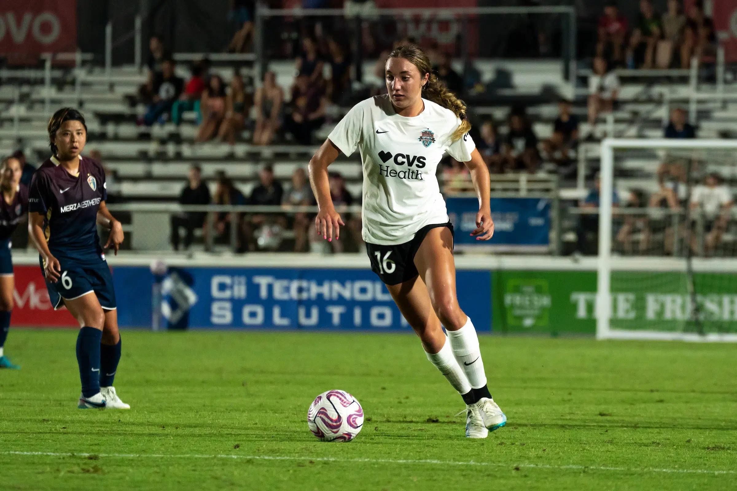 Nicole Douglas in a white top, black shorts and white socks dribbles a soccer ball as a defender in a dark uniform looks on.