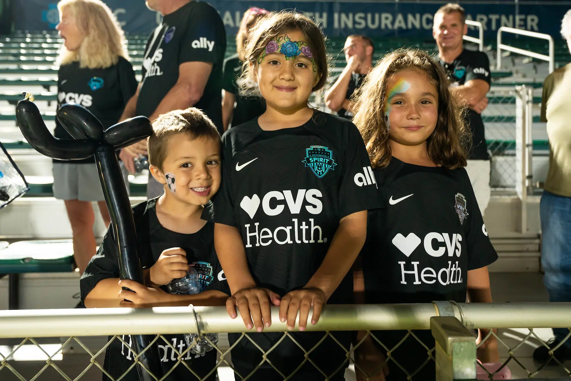 A young boy and two girls with decorative and sparkly face paint wear matching black uniforms and stand along the bleachers of a soccer stadium.
