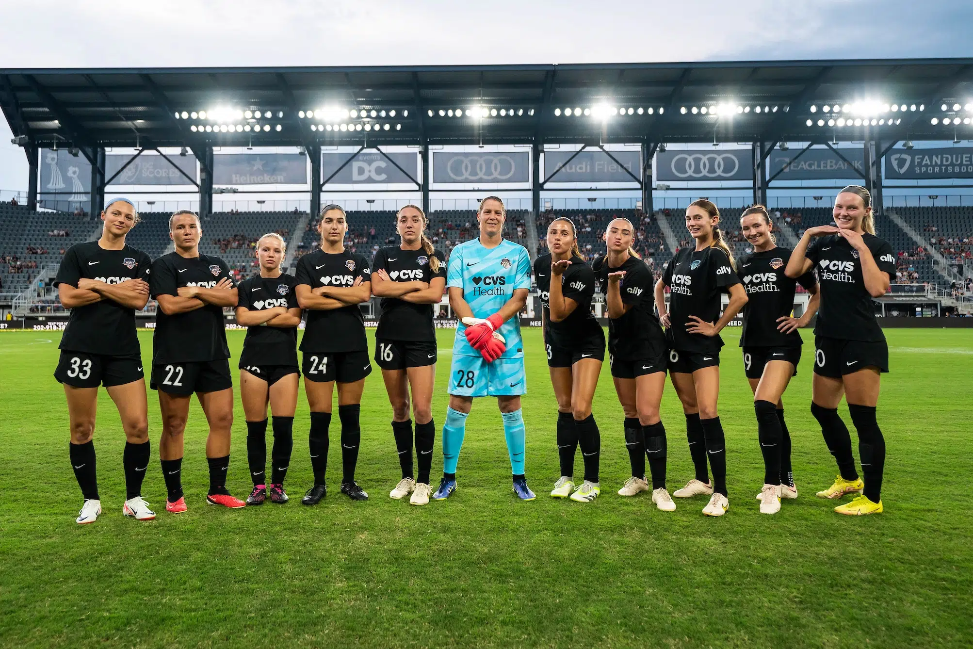 On the left side, Five soccer players in black uniforms stand with their arms crossed and have serious looks on their faces. On the right side, five more players in black blow kisses and pose with their hands on their hips smiling wide. In between the two groups, Nicole Barnhart wears a blue goalie uniform and red gloves.