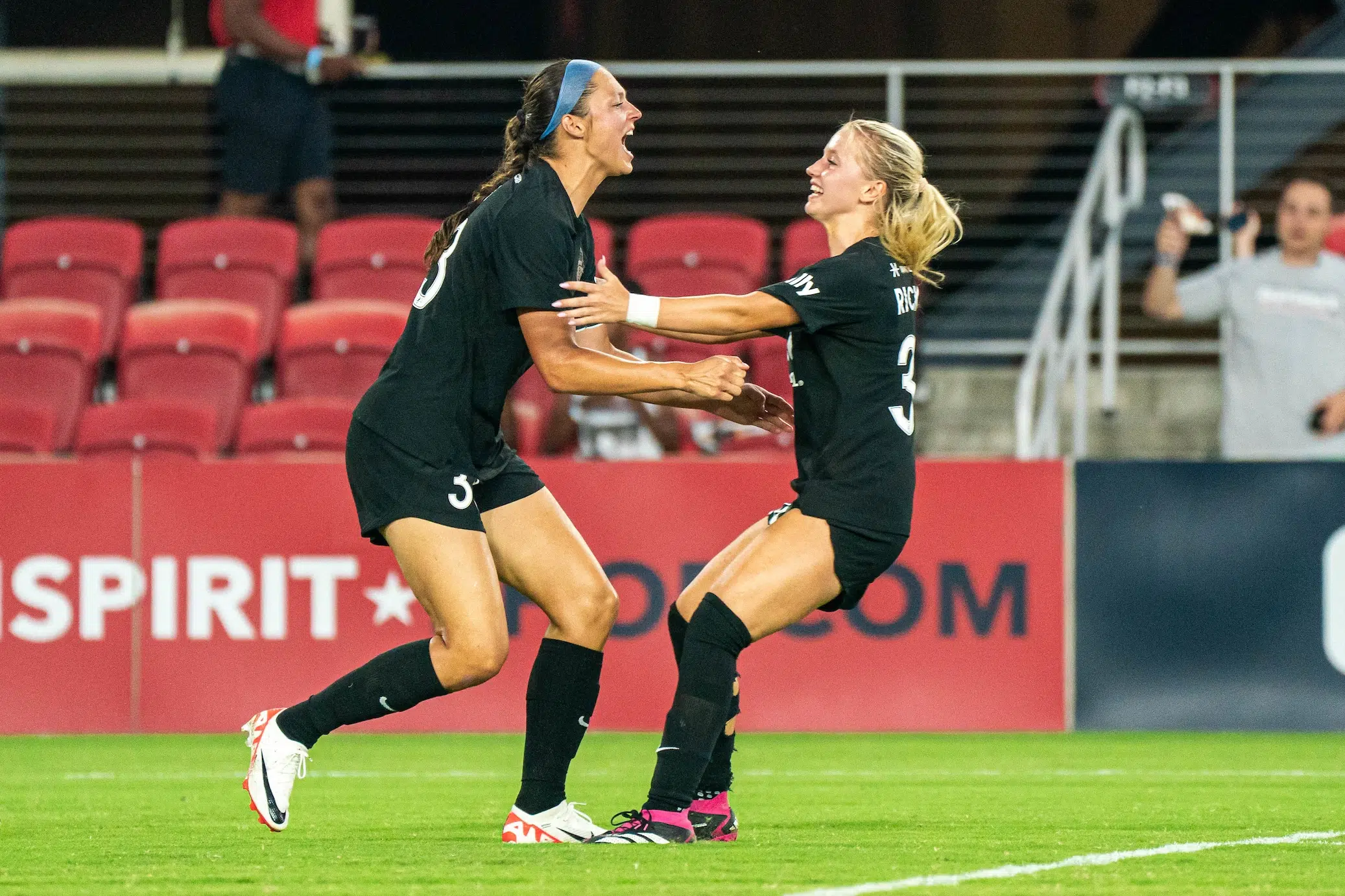 Ashley Hatch and Chloe Ricketts, both in black uniforms, extend their arms and are about to embrace on a soccer field.
