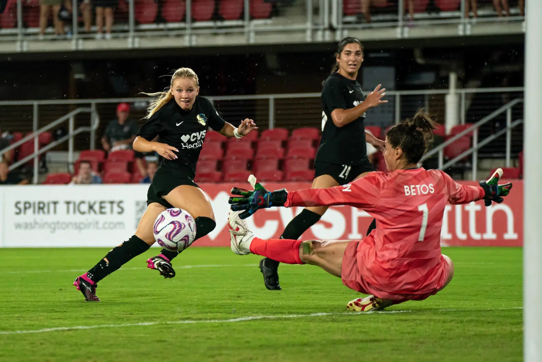 Chloe Ricketts in a black uniform watches the soccer ball she just shot as a goalie in a red uniform attempts to save it.
