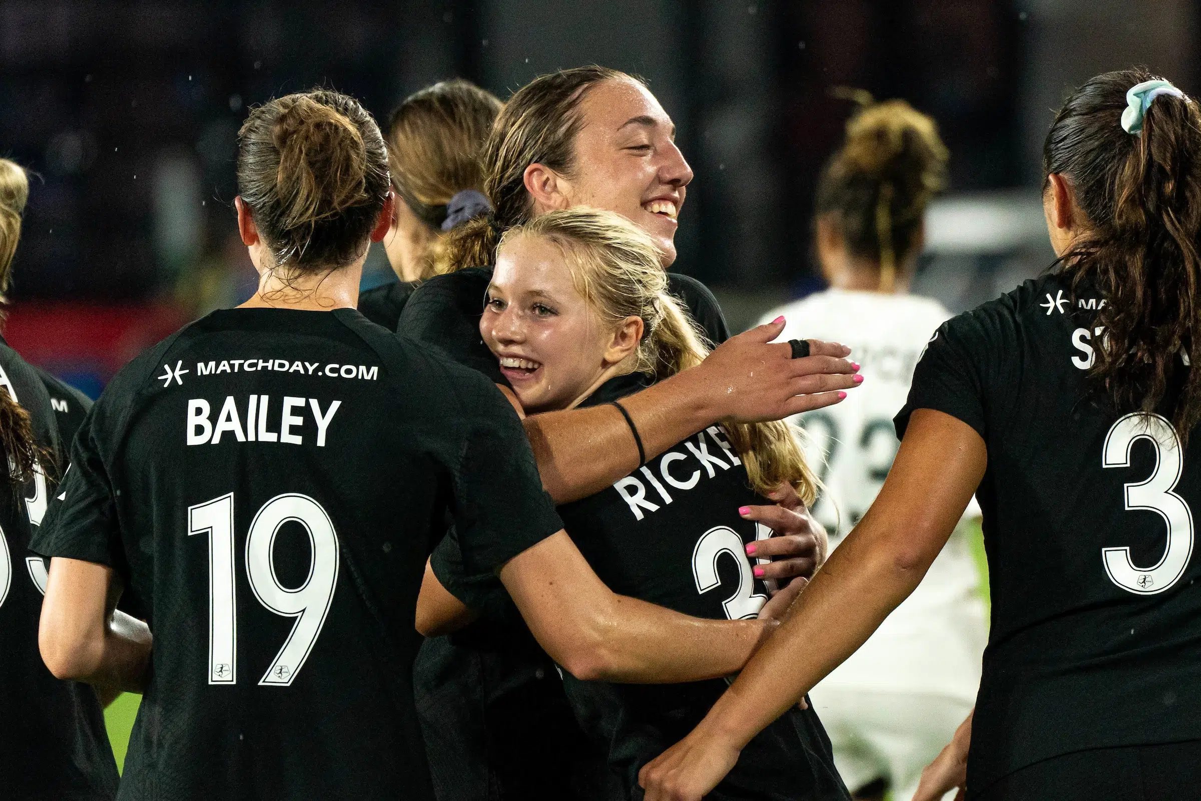 A group of soccer players in black uniforms hug Chloe Ricketts who smiles.