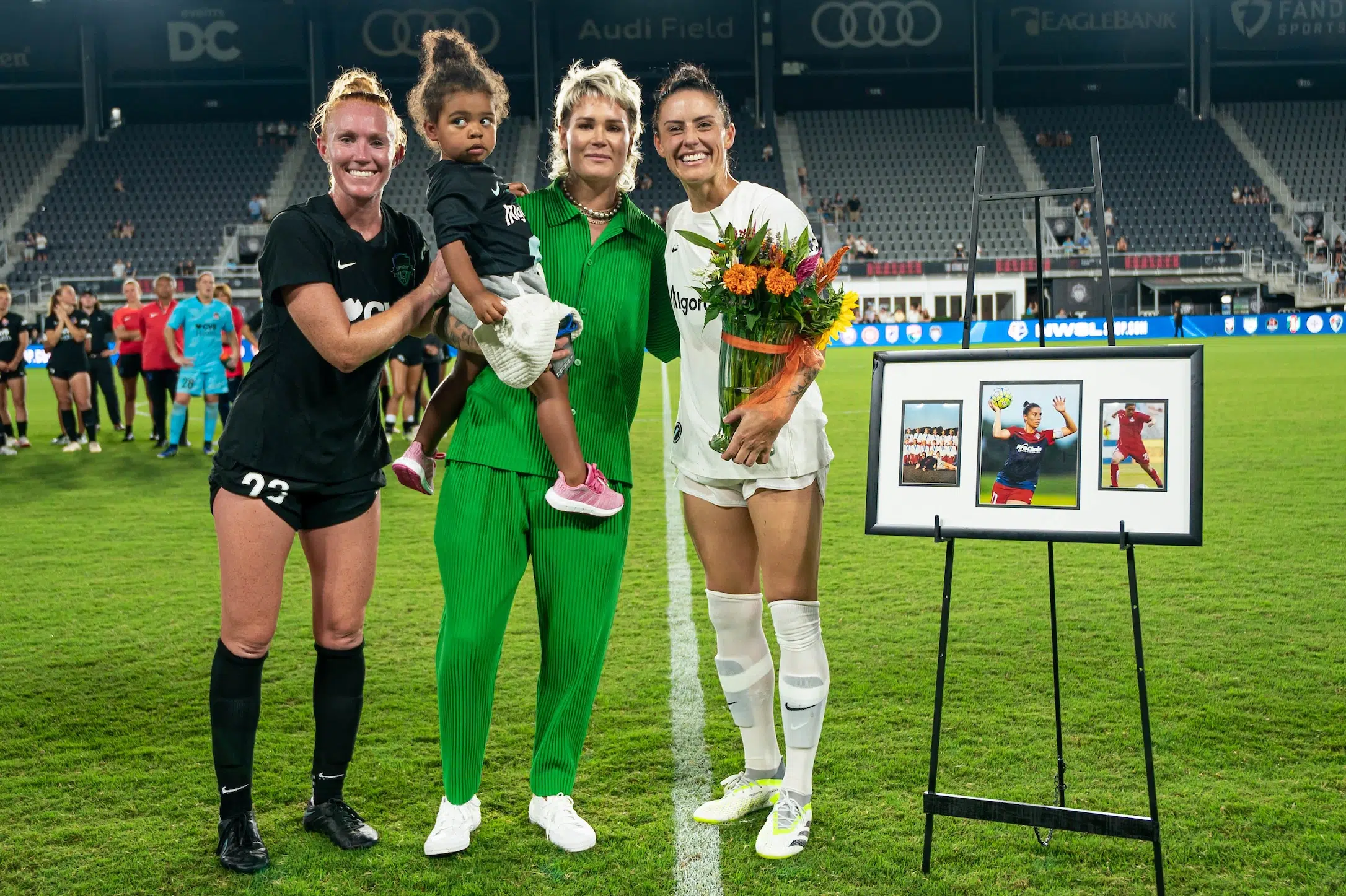 Tori Huster in a black soccer uniform, Ashley Harris in matching green pants and shirts, and Ali Krieger in a white uniform holding a bouquet of flowers smile next to an easel featuring a picture frame with three pictures of Krieger. Harris holds a child in a black shirt and pink shoes.