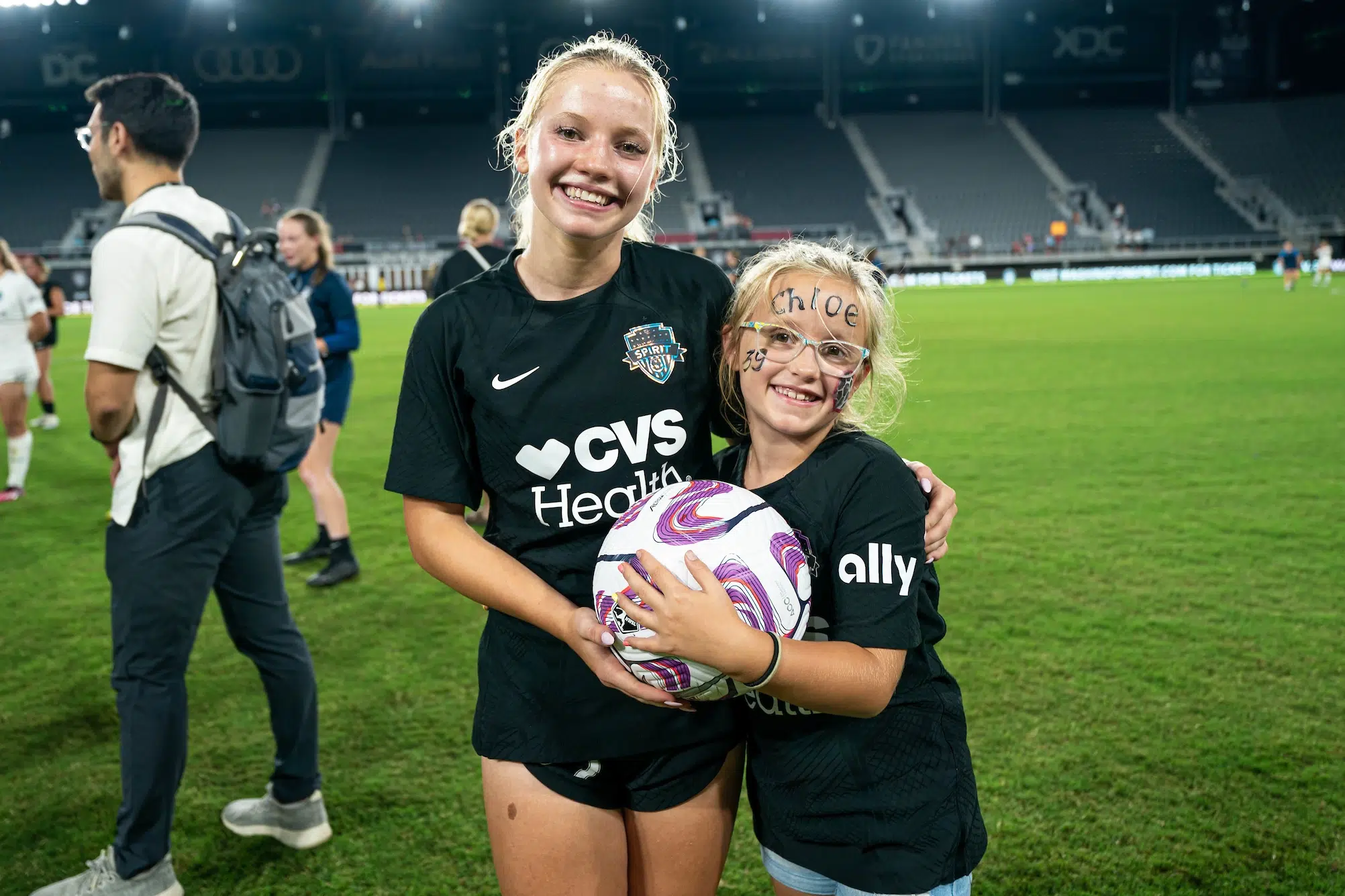 Chloe Ricketts in an all black uniform side hugs a younger child in a Ricketts jersey as the two hold a soccer ball together.