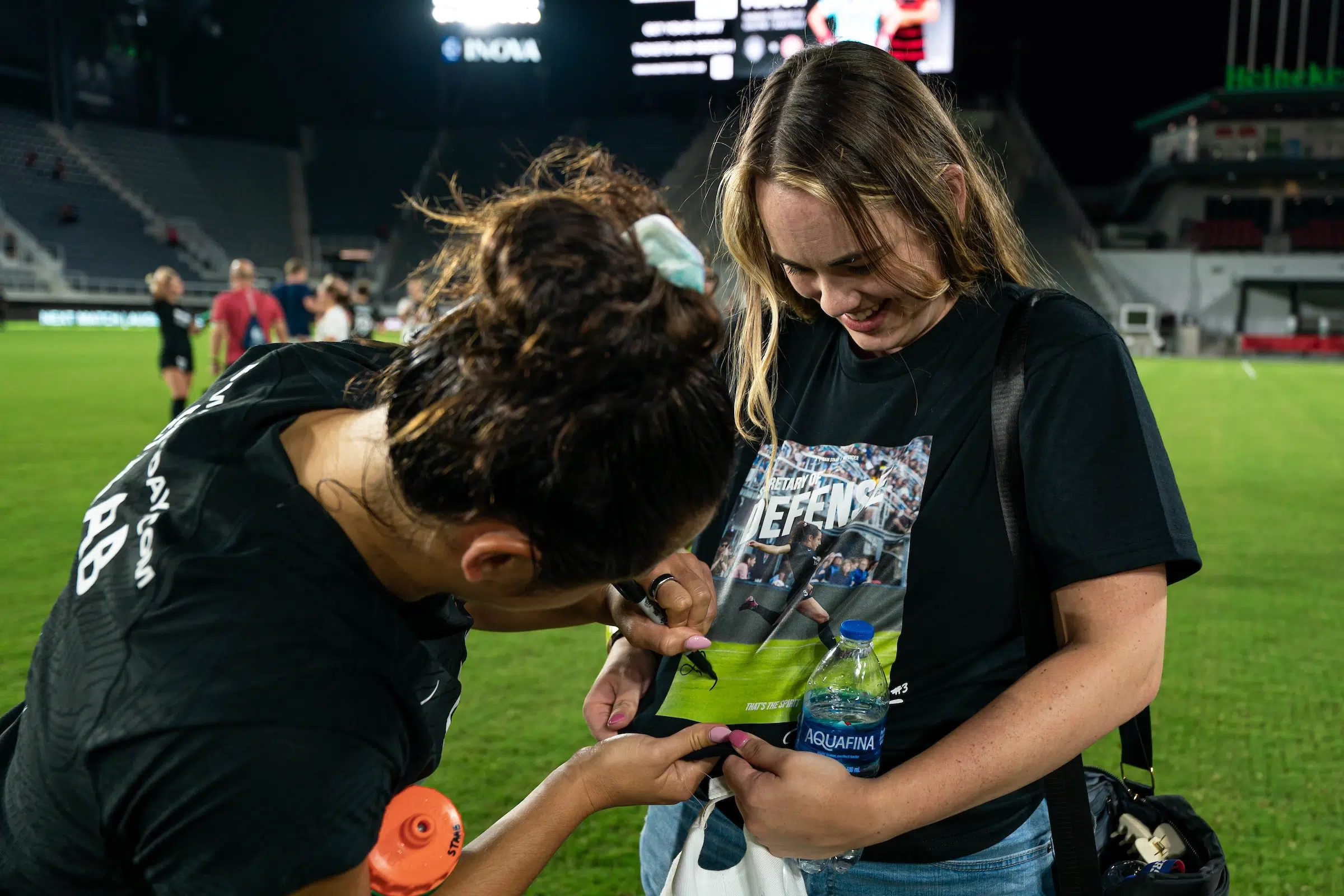 Sam Staab signs a fan's t-shirt which features an image of Staab. The fan is holding the shirt away from her body and looking down as Staab signs.