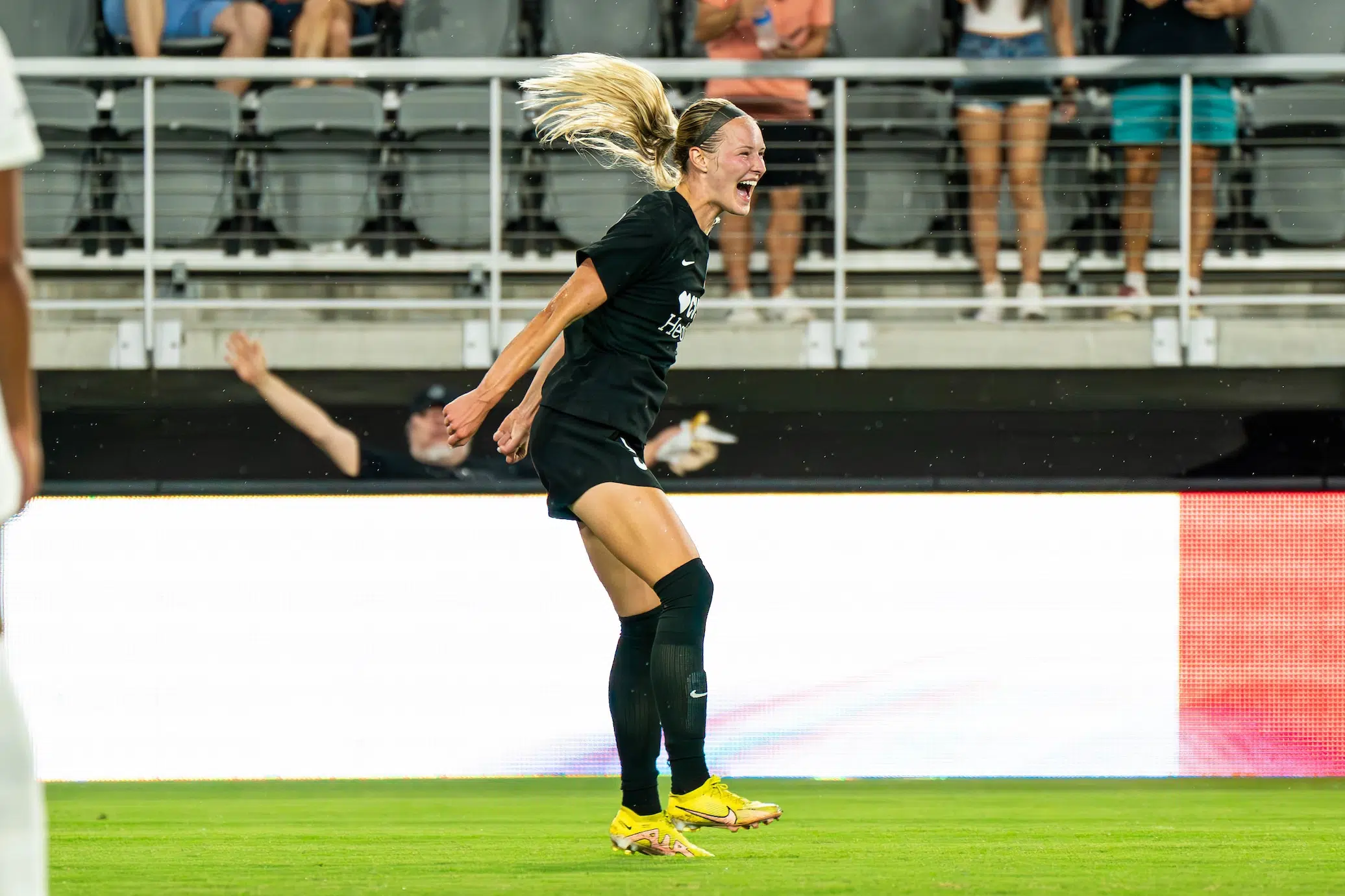 A soccer player in a black uniform jumps up as her blonde ponytail flies in the air. She has a big smile on her face and is wearing yellow cleats.
