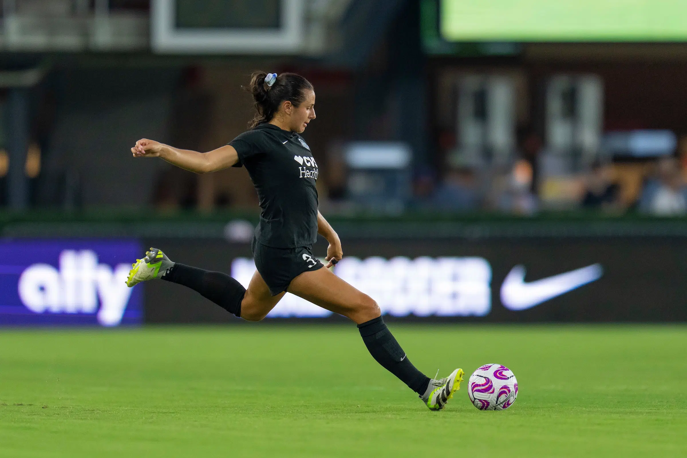 Sam Staab in an all black uniform with her hair in a pony tail with a scrunchie, winds up to kick a soccer ball on a green field.