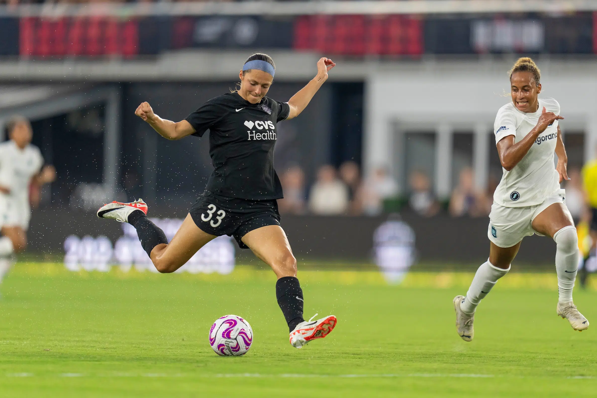 Ashley Hatch in a black uniform and white and red cleats, winds up to kick a soccer ball as a defender in an all white uniforms tries to keep up.