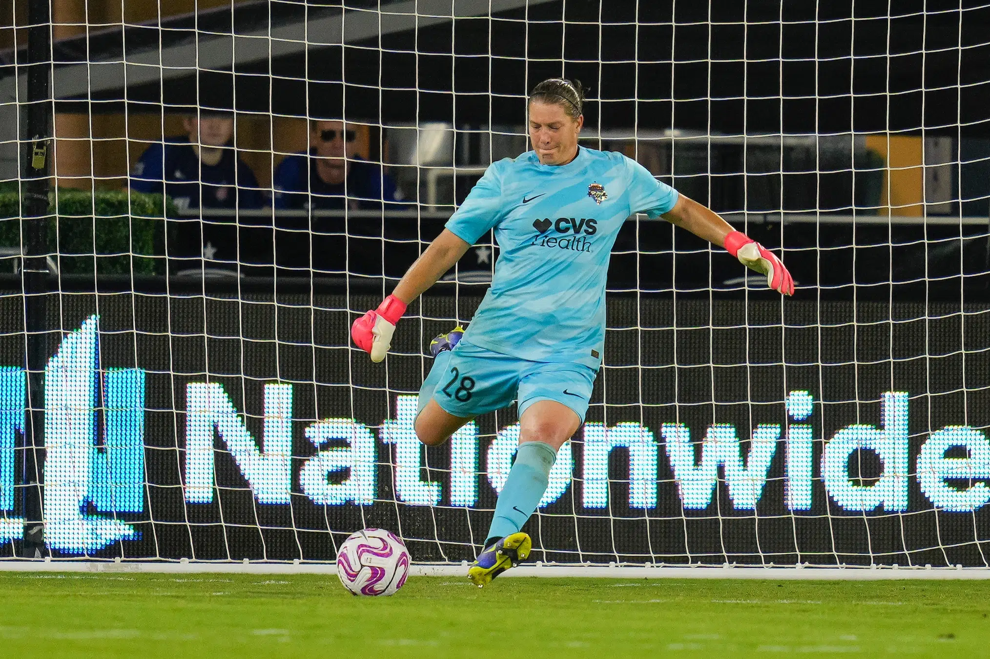 A goalie in a blue uniform winds up to kick a soccer ball. The net is visible in the background.