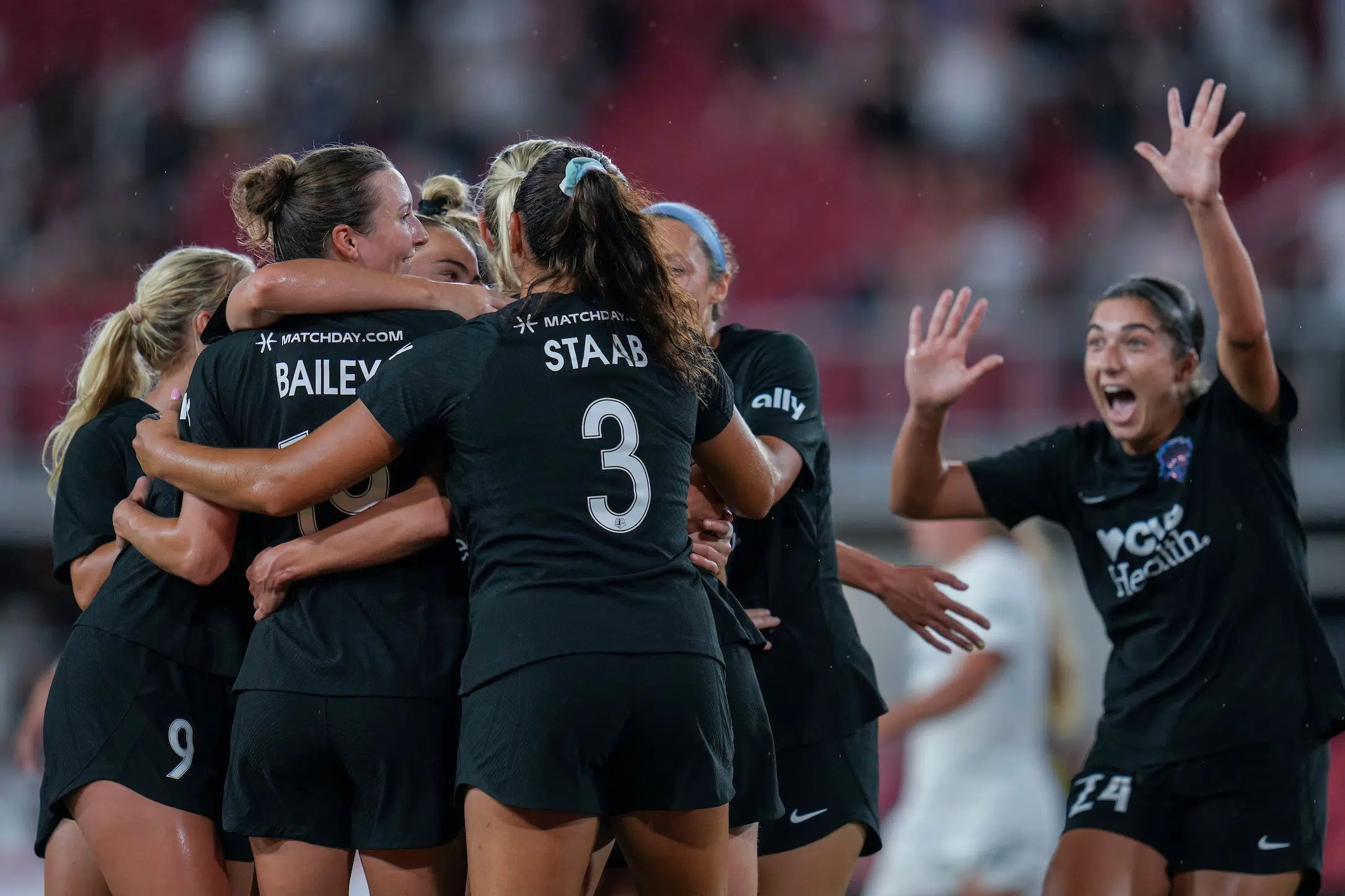 A group of soccer players in black uniforms hugs in celebration as Lena Silano in a black uniform runs towards the group with a wide smile and hands in the air.
