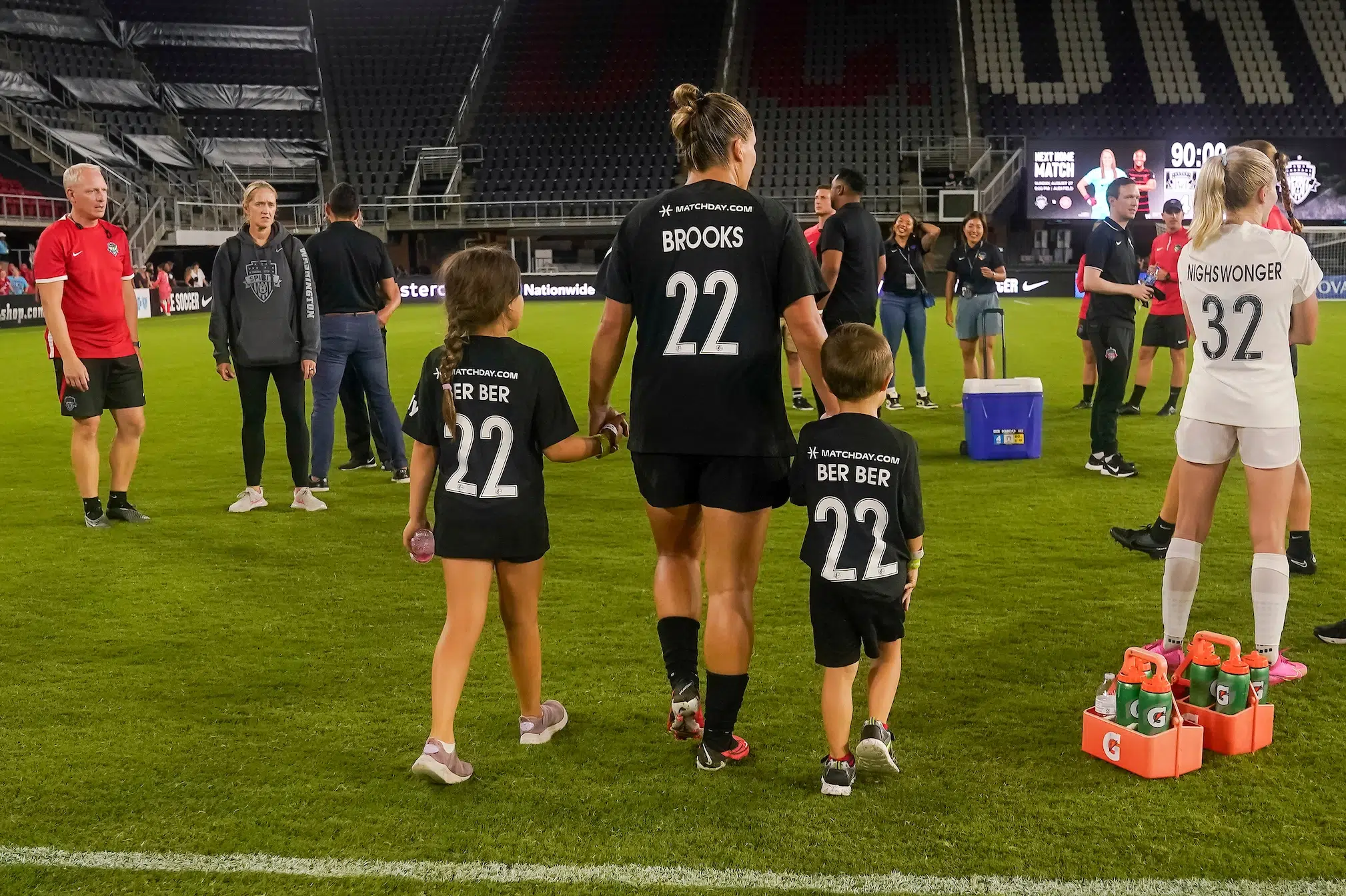 Dressed in all black uniforms, Amber Brooks walks away from the camera on the soccer pitch at Audi Field holding hands with a young girl and young boy. They're both wearing replicas of her black jersey.