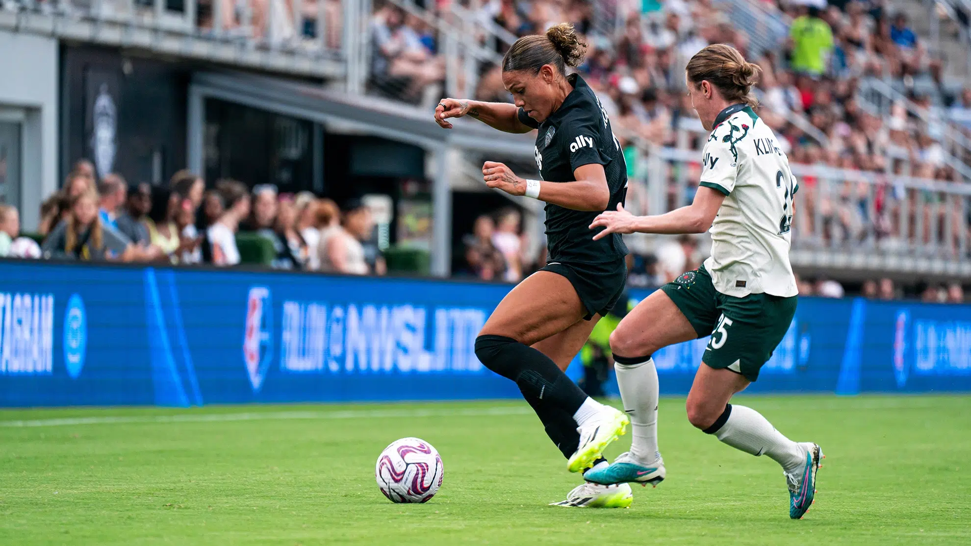Trinity Rodman in an all black uniform dribbles the ball and works to keep it away from a defender in a white top, green shorts and white socks.