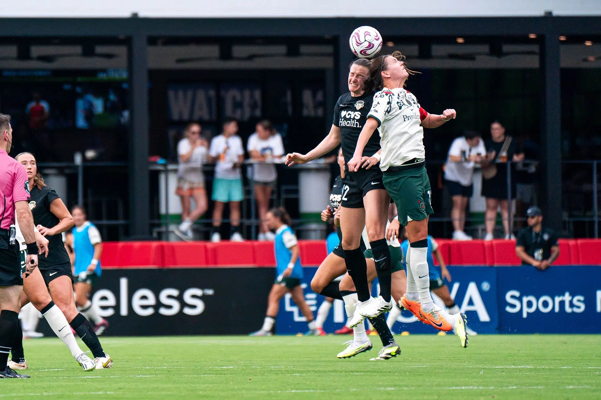 Andi Sullivan in a black uniform jumps up to head the ball as Christine Sinclair in a white top and green shorts tries to challenge her.