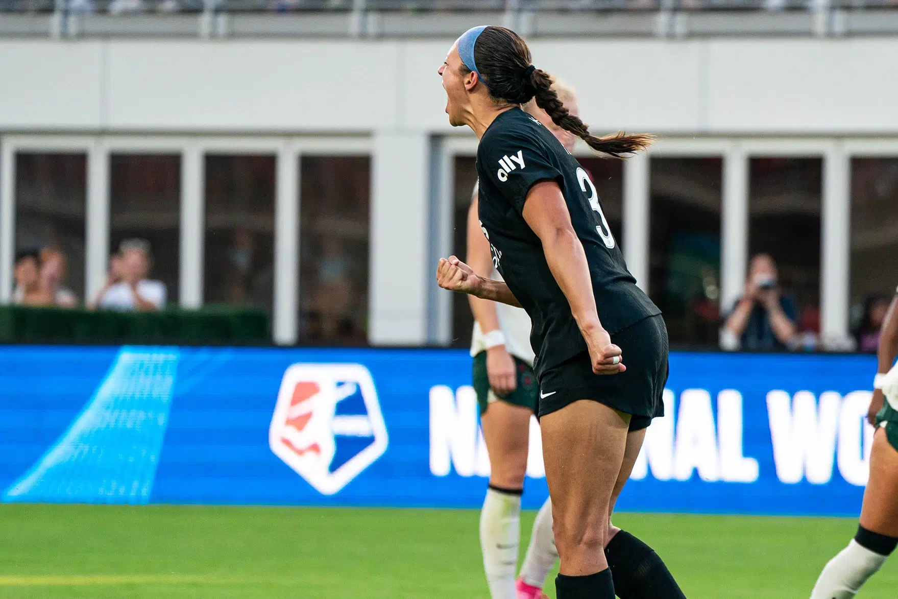 Ashley Hatch in an all black uniform with her hair in a braid pumps her fist and yells in celebration of scoring a penalty kick.