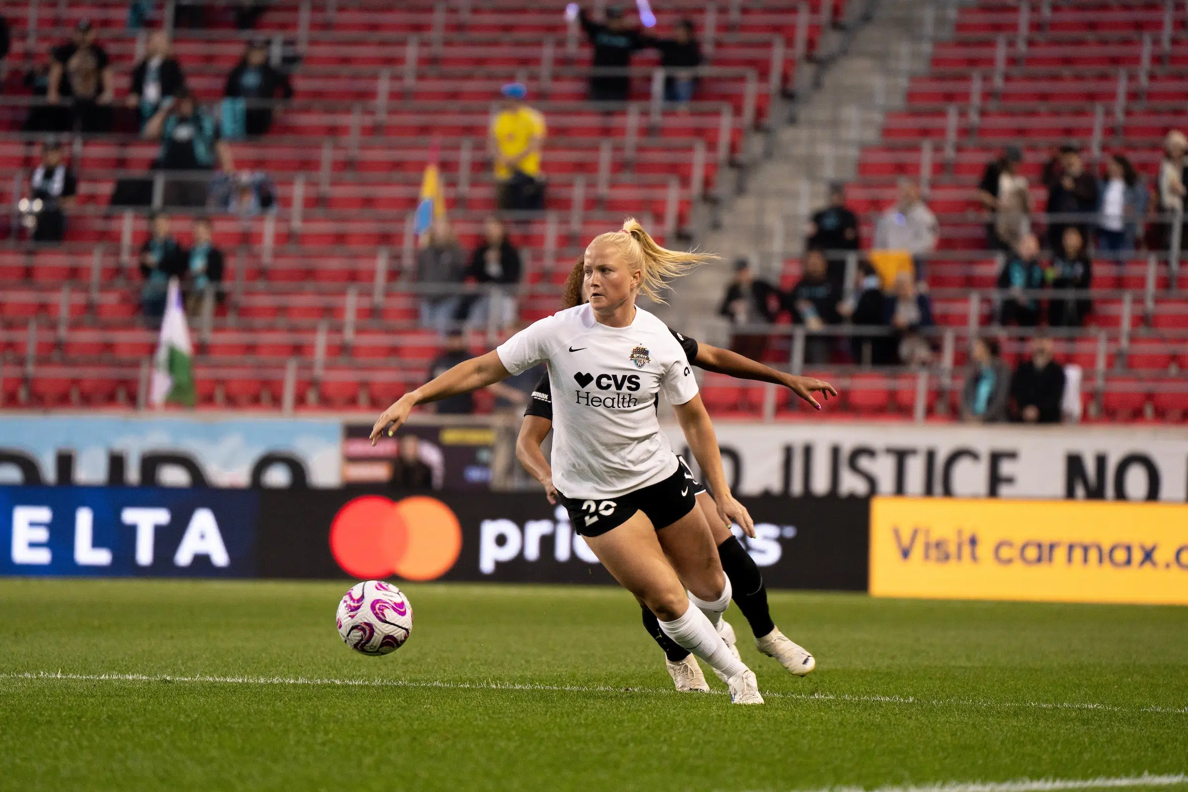 Civana Kuhlmann looks forward as she dribbles a soccer ball. She is wearing a white top, black shorts and white socks. A defender is barely visible as she tries to chase Kuhlmann down.