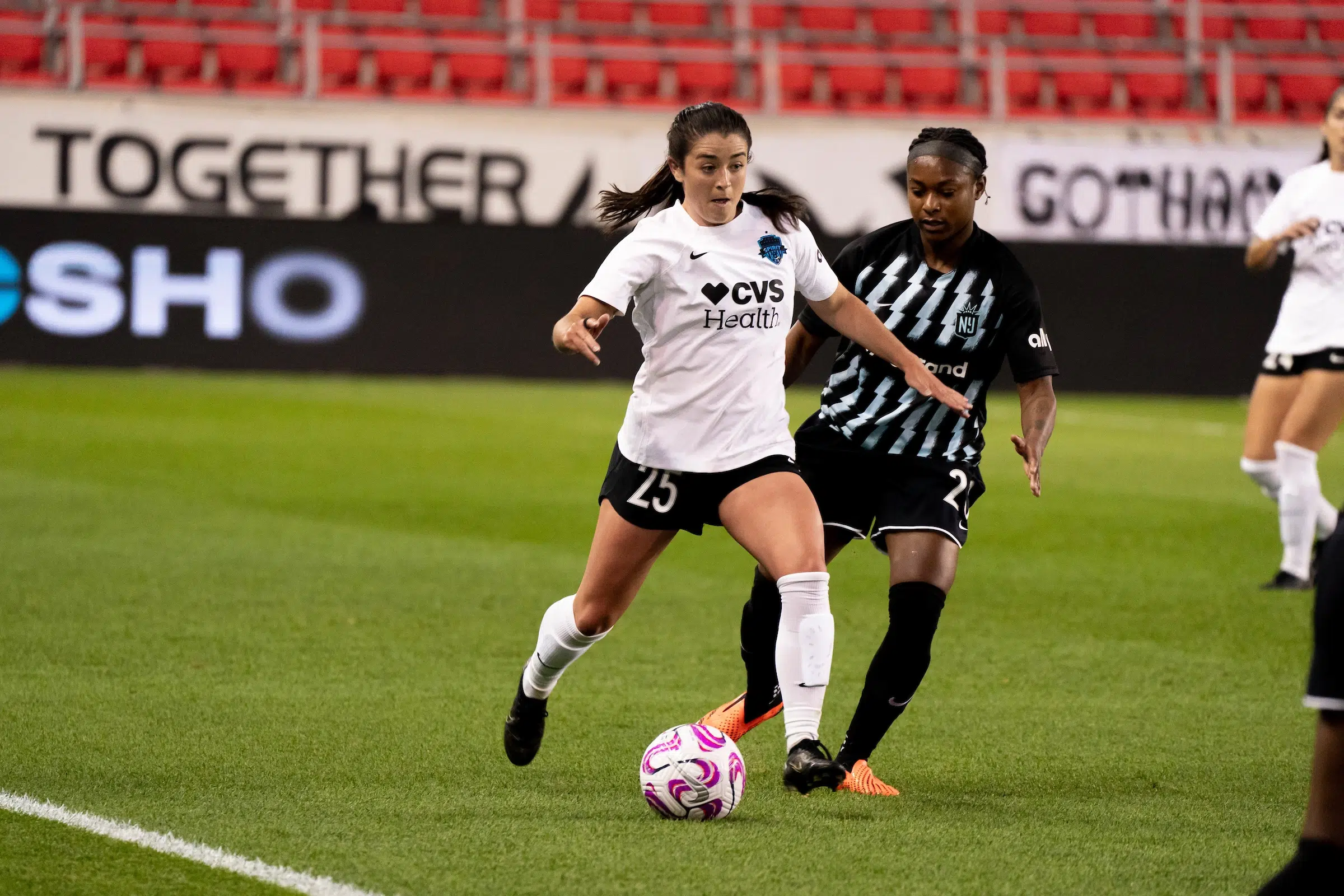 Marissa Sheva in a white top, black shorts and white socks dribbles a soccer ball as a defender in a black uniform chases her down.