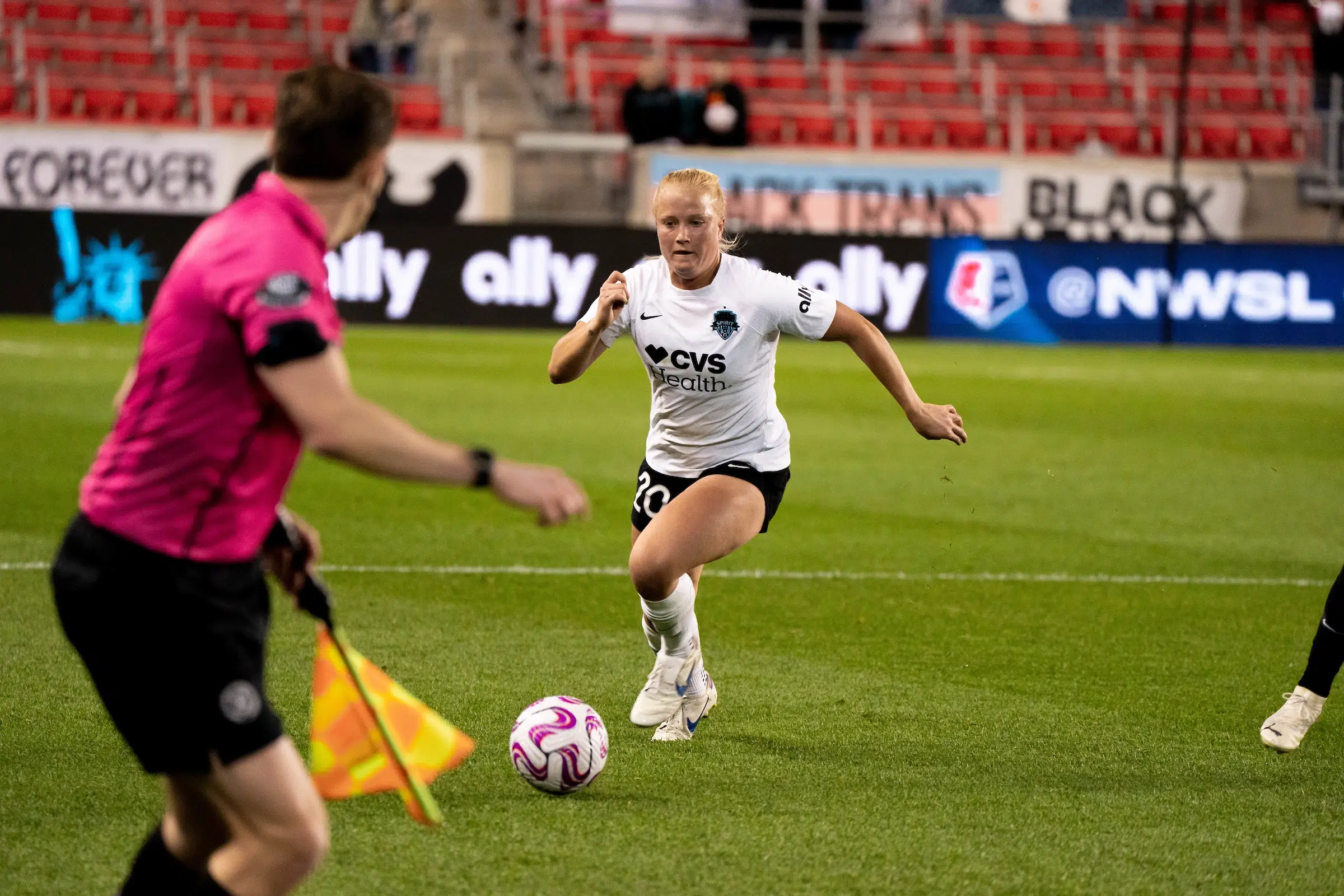 Civana Kuhlmann dribbles a soccer ball. In the foreground, a soccer referee in a pink shirt is out of focus.