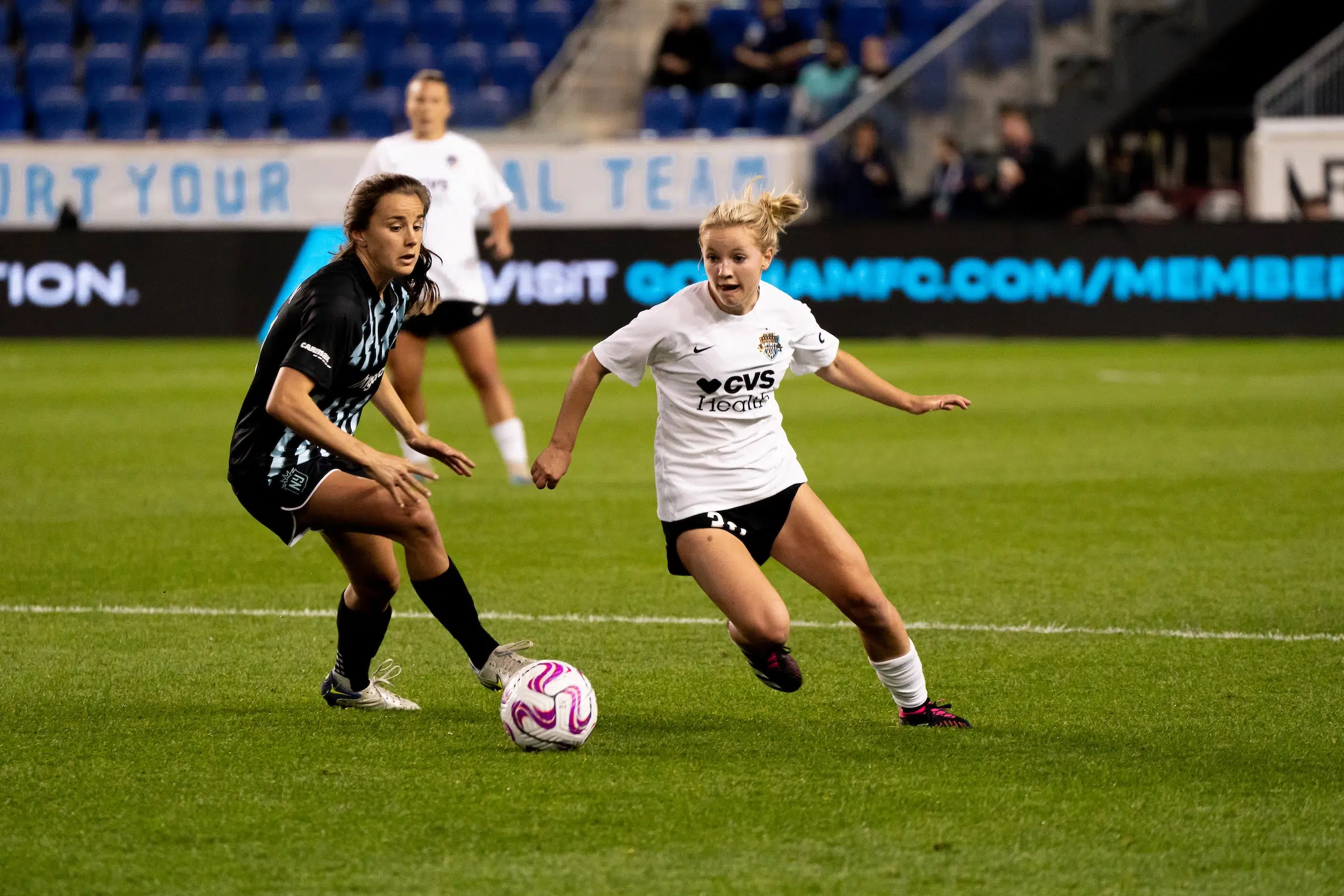 Chloe Ricketts in a white top, black shorts and white socks dribbles past a defender wearing a black uniform.