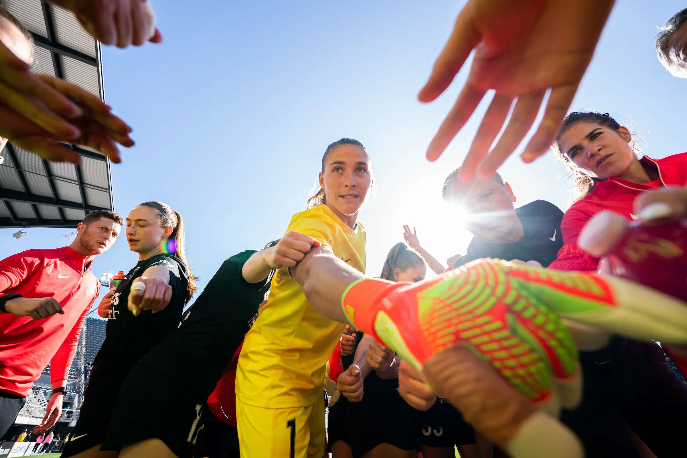 Aubrey Kingsbury in a yellow goalie uniform and neon orange and green gloves puts her hand in a huddle of soccer players.