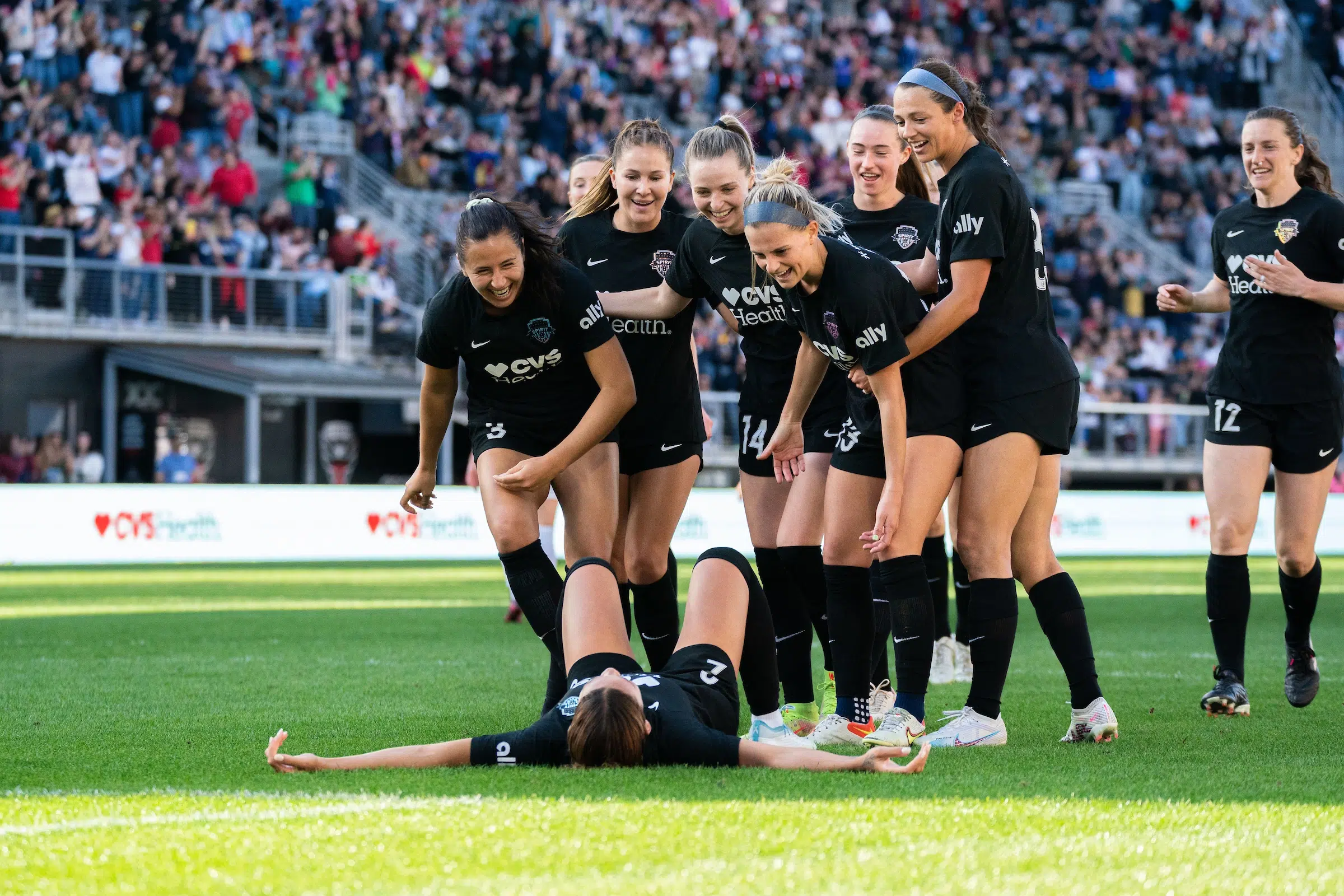 A group of soccer players in black uniforms smile and look down at their teammate who is sprawled on the ground laughing in celebration of a goal.