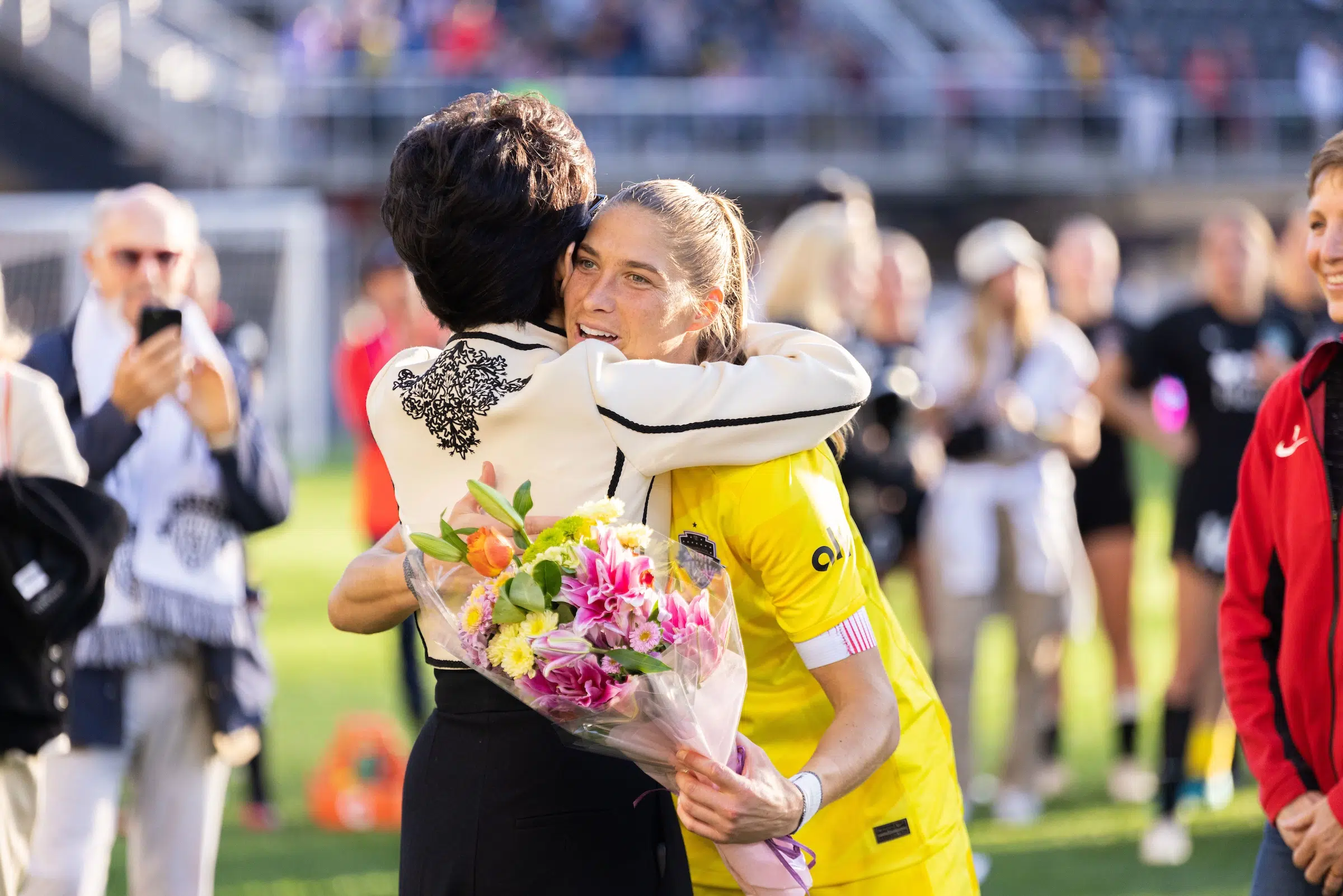 Aubrey Kingsbury in a yellow uniform holds a bouquet of flowers in one hand and hugs Michele Kang.
