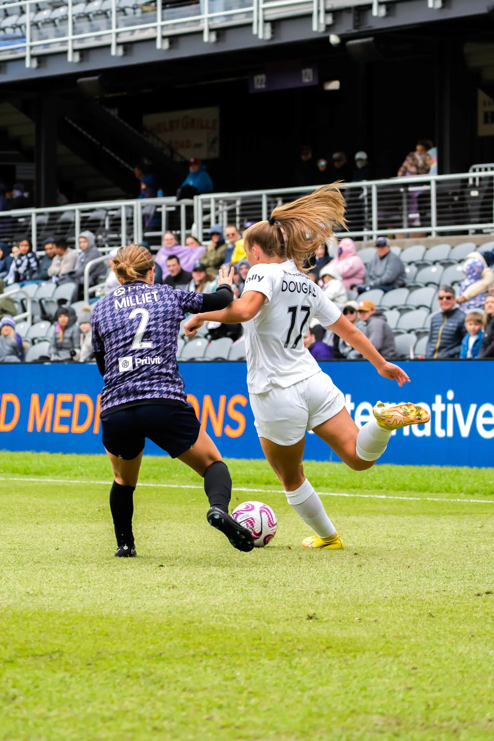 Two soccer players, one in an all-white uniform and one in a purple-and-black houndstooth uniform, battle for possession of a soccer ball.
