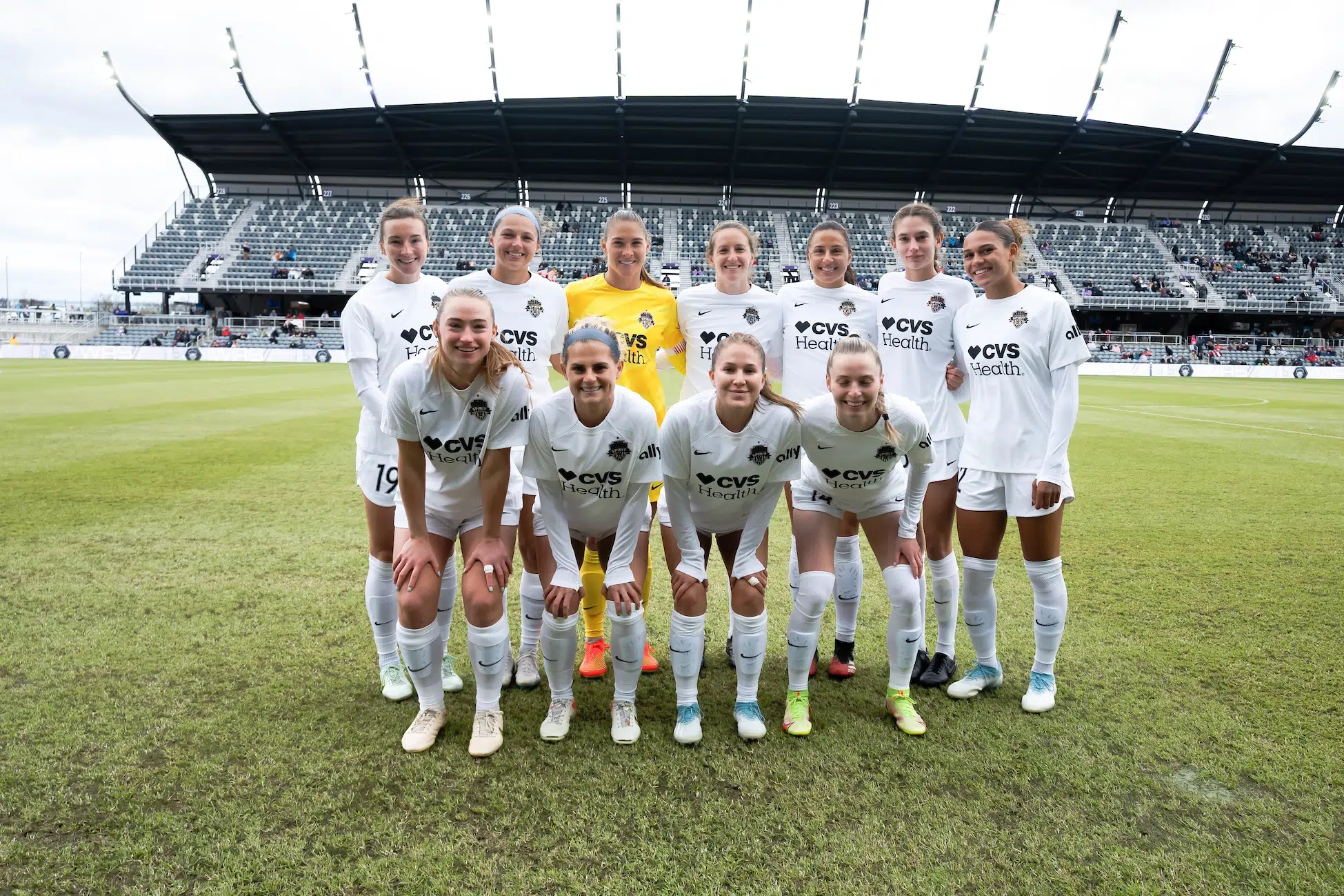 Ten players in all-white uniforms and one in a yellow goalie uniform pose for a photo in two rows on a soccer field.